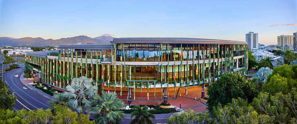 The Cairns Convention Centre, a modern multi-story building with vertical green features and large glass windows, surrounded by lush greenery and trees, with mountains in the background and a clear blue sky.