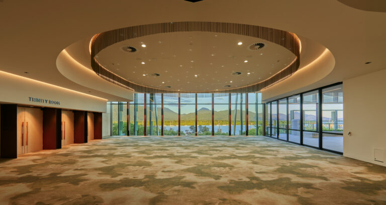 The Trinity Room at the Cairns Convention Centre is a spacious, modern venue featuring a large circular ceiling light fixture with recessed lighting and spotlights. The floor is covered in a patterned carpet of brown and beige tones. Three wooden doors on the left are labeled 'TRINITY ROOM.' Floor-to-ceiling windows at the far end offer a scenic view of lush greenery and distant mountains, allowing natural light to fill the room