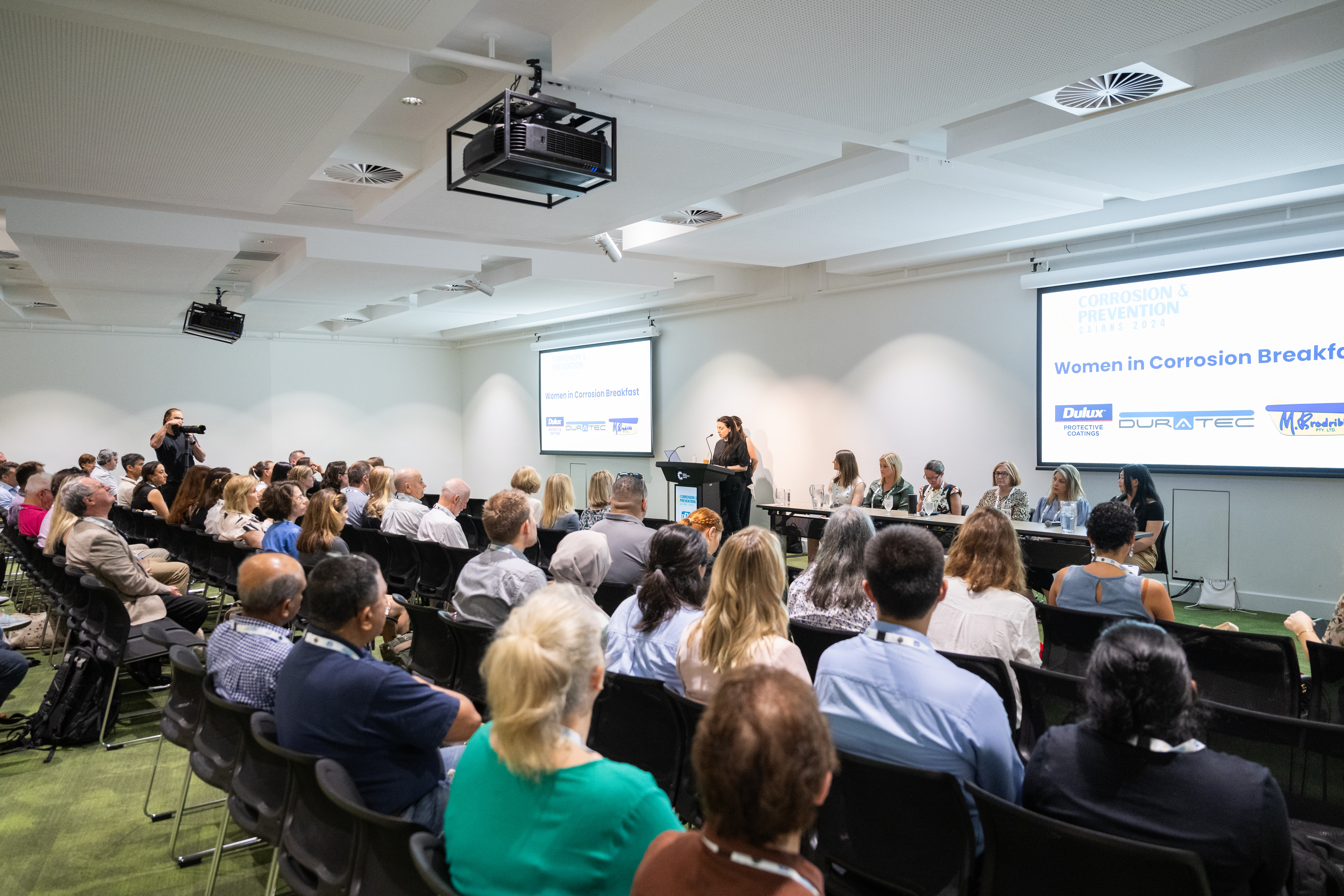 The image is situated in meeting rooms of the Cairns Convention Centre, with an audience seated and facing a panel of speakers at the front. The event is titled 'Women in Corrosion Breakfast,' as shown on the projection screen. A speaker stands at a podium addressing the audience. The room features white walls, ceiling lights, and ceiling-mounted projectors