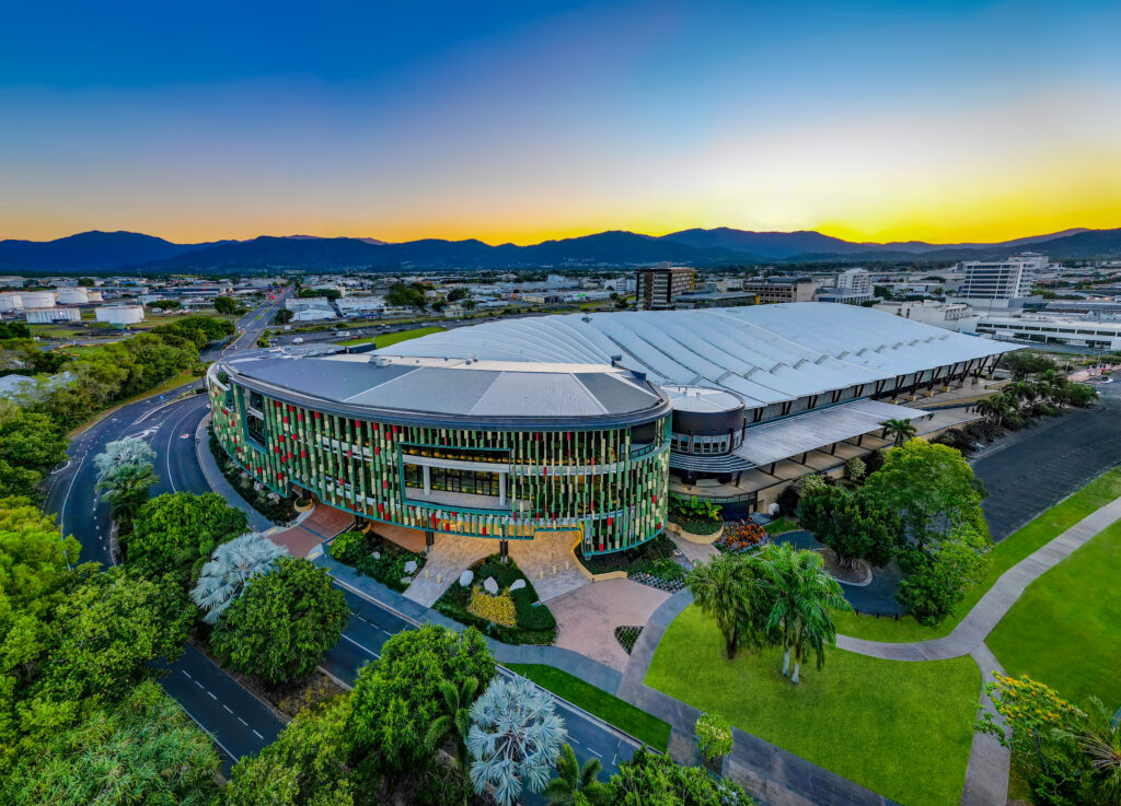 Aerial view of the Cairns Convention Centre, featuring a modern circular building with a colorful facade, surrounded by greenery and roads, with a cityscape, mountains, and a vibrant sunset in the background