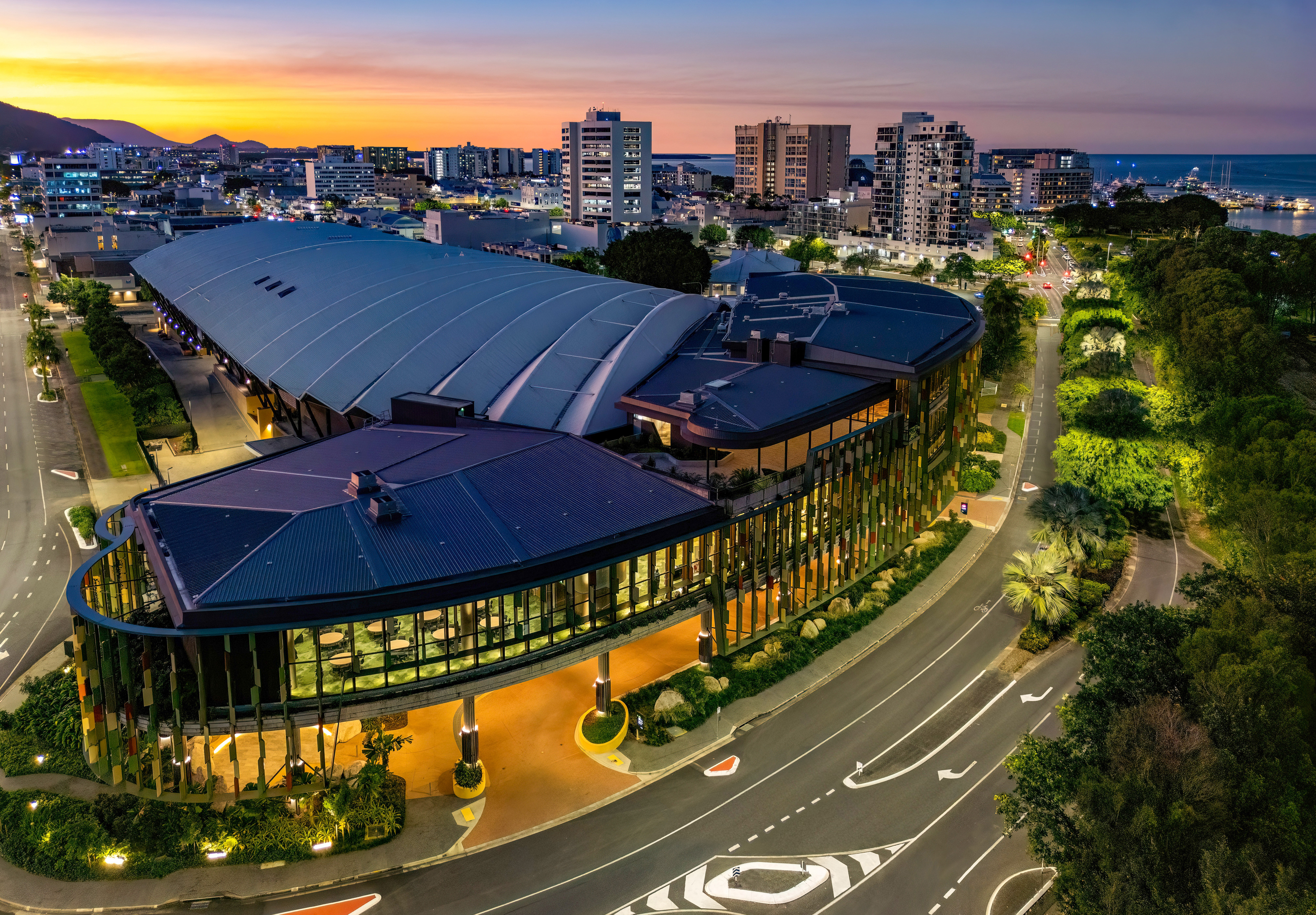 Aerial view of the Cairns Convention Centre with its curved, glass-accented architecture, surrounded by roads and greenery, with city buildings and the sea visible at sunset