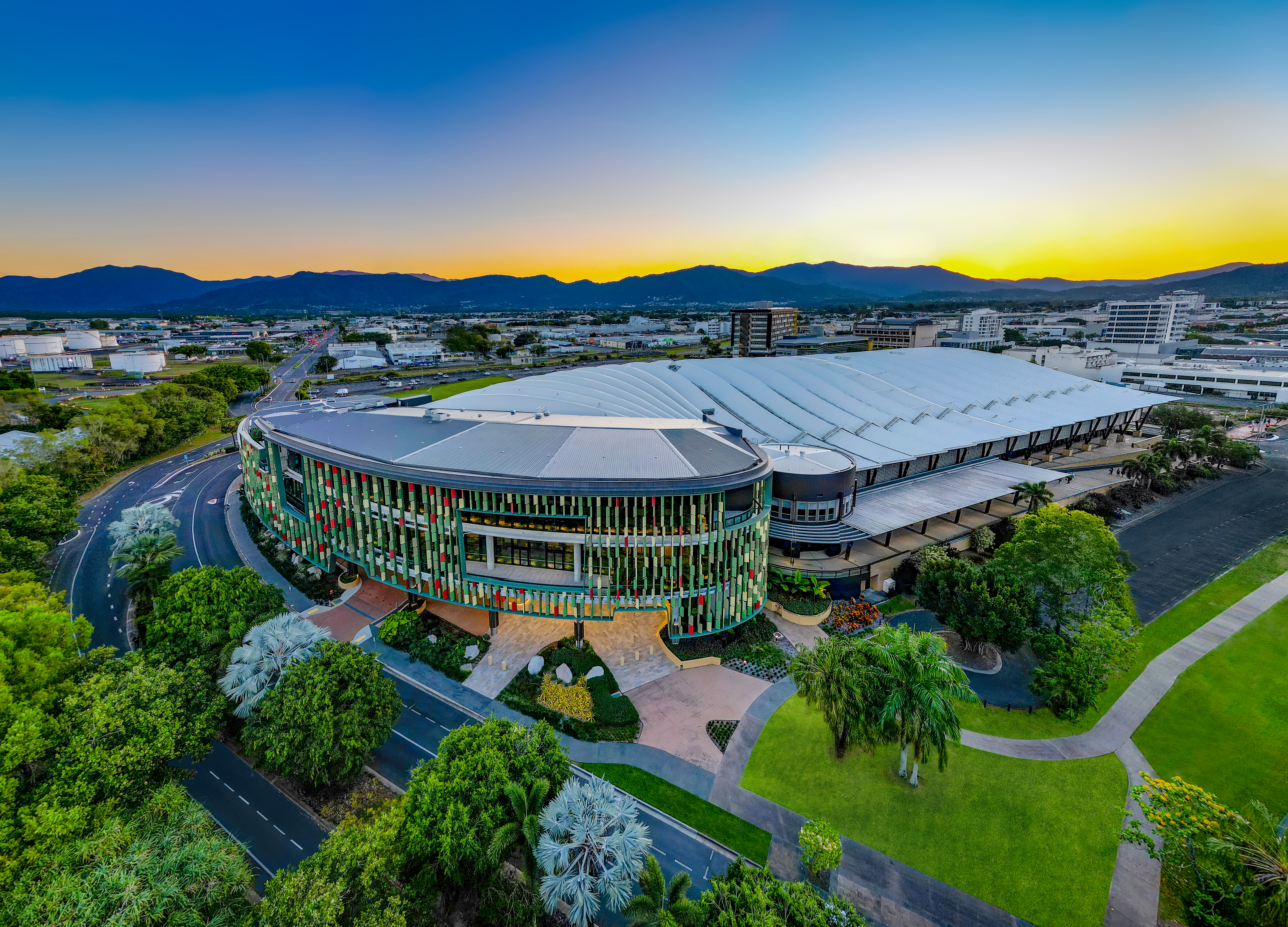 Aerial view of the Cairns Convention Centre, featuring a modern circular building with a colorful facade, surrounded by greenery and roads, with a cityscape, mountains, and a vibrant sunset in the background