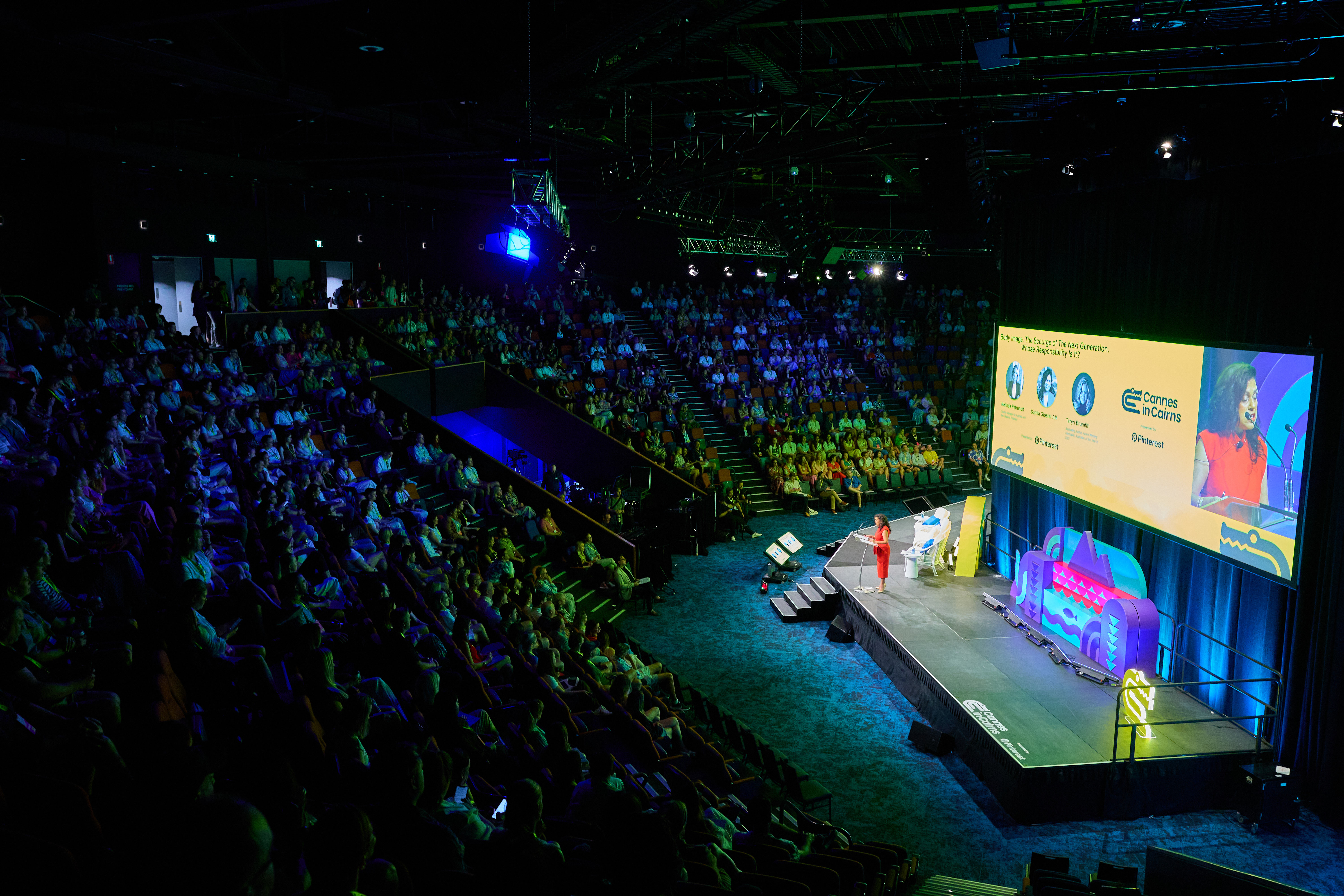 A large auditorium filled with people attending a presentation or conference. A speaker in red attire is presenting on a brightly lit stage at the front, which features multiple screens displaying images and text. The audience is seated in rows, and the rest of the room is dimly lit