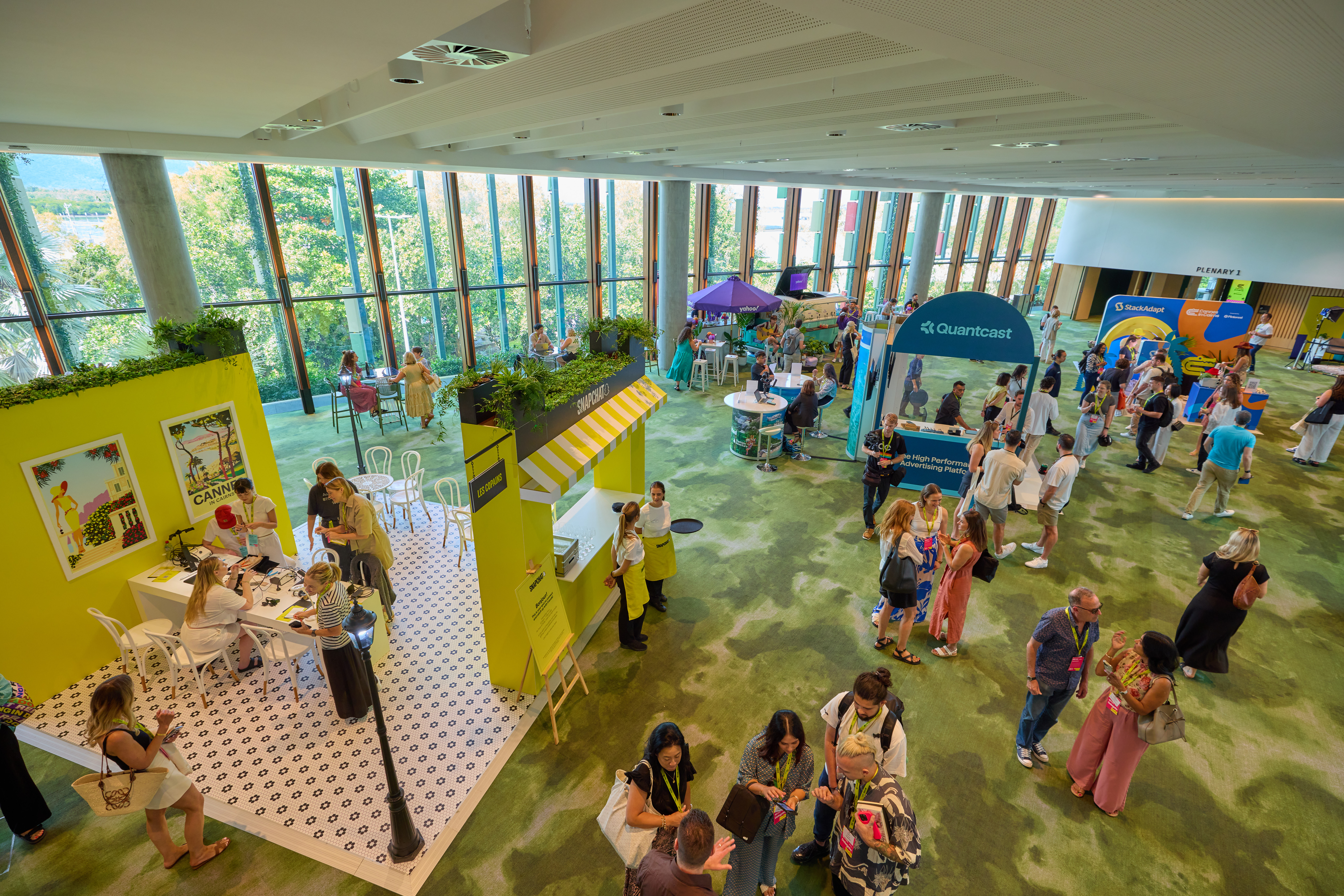 A large indoor event space on the mezzanine level of the Cairns Convention Centre, featuring several booths and people interacting. The room has large windows letting in natural light and a green carpeted floor. Booths include one with a yellow and white striped awning, another labeled 'Claimrnest,' and others in the background. Attendees are walking, talking, and engaging with booth representatives in a lively, busy atmosphere
