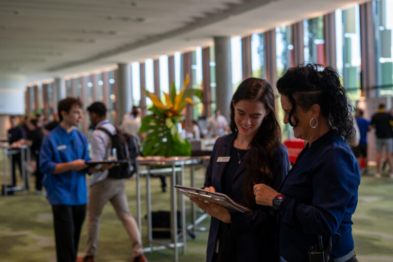 A group of people in a large, well-lit room with floor-to-ceiling windows. Two individuals in the foreground hold clipboards and wear name tags. In the background, others are standing and conversing around high tables. The room features modern design elements, green carpeting, and decorative plants