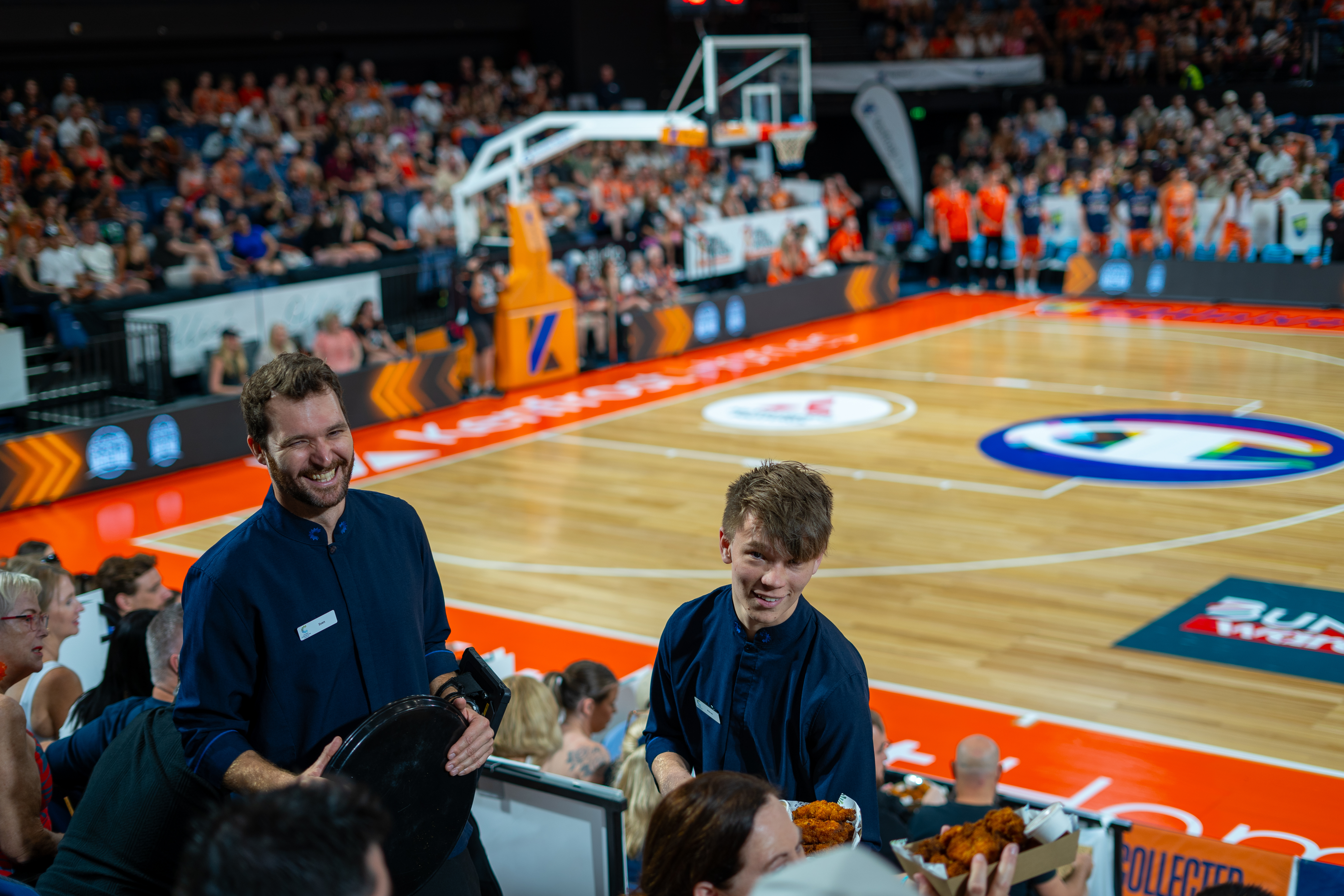Two Cairns Convention Centre servers in navy blue uniforms are seen standing in front of a crowded basketball arena. The court is brightly lit with orange and white markings, and the stands are filled with spectators. The servers appear to be attending to the audience seated near the court
