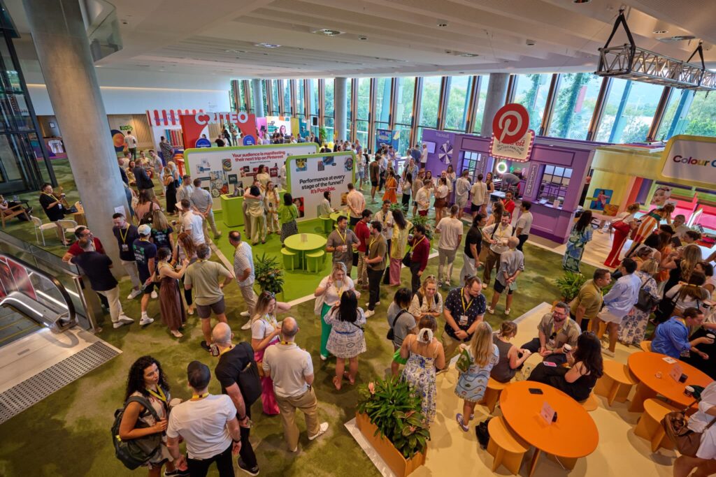 A lively mezzanine exhibition space at the Cairns Convention Centre, filled with attendees engaging in conversations and exploring colorful booths. The area is well-lit with natural light streaming through large windows. Several round tables with green and orange tops and decorative plants are scattered throughout the space. An escalator is visible on the left side, and prominent signage for Pinterest and other brands is displayed among the booths