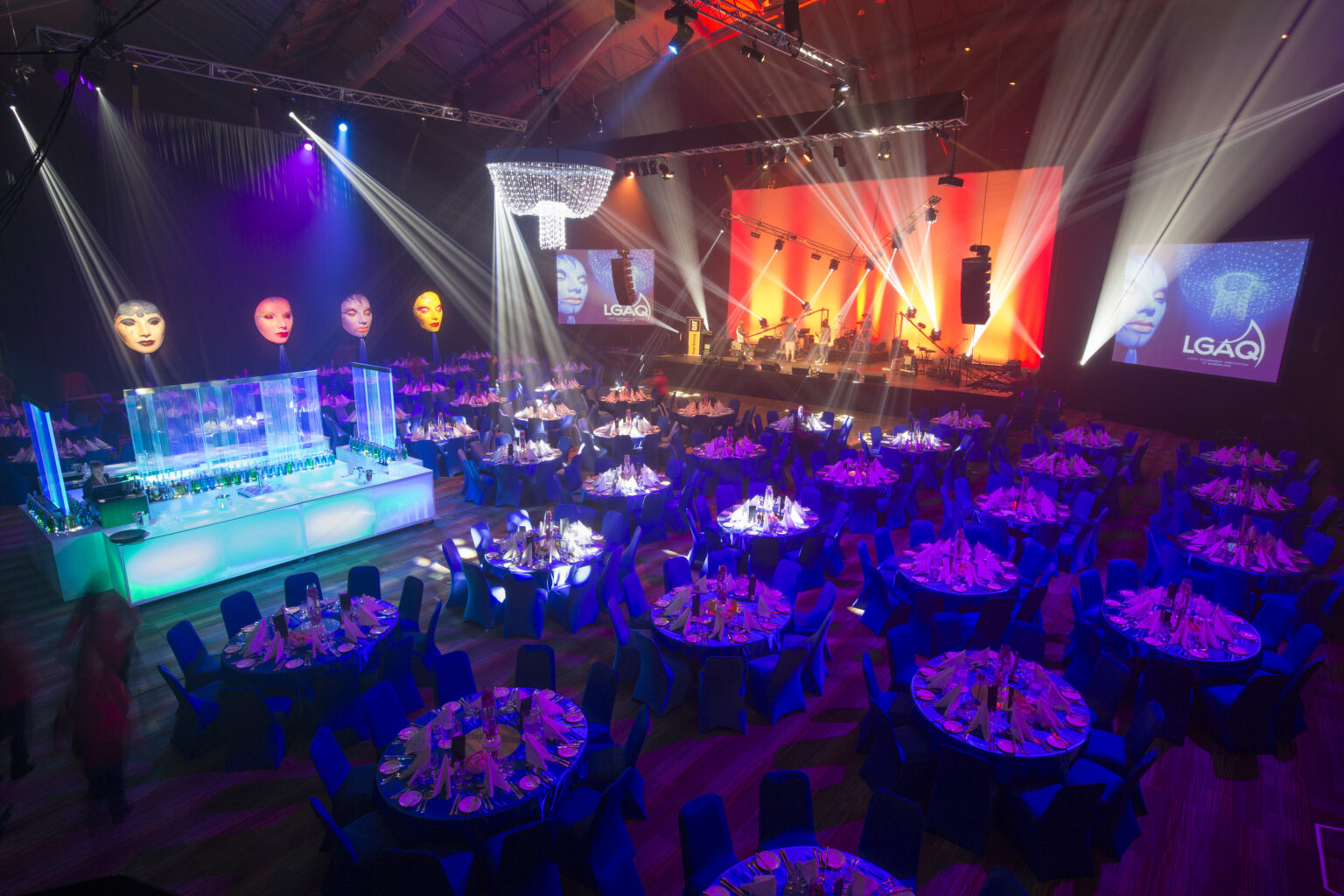 A gala dinner event in the arena at the Cairns Convention Centre, featuring round tables with blue tablecloths, elegantly set with plates, glasses, and napkins. The space is illuminated by colorful lighting and spotlights, creating a vibrant atmosphere. A stage at the far end displays a large screen with 'LGAQ' and images of faces. Decorative elements, including large masks and a chandelier-like structure, enhance the festive ambiance