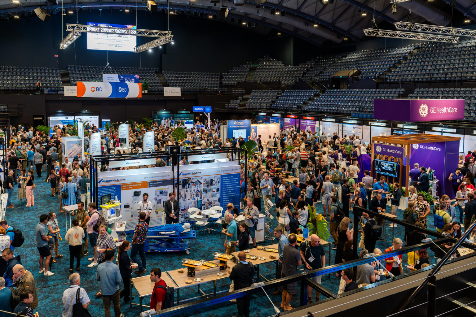 A bustling photo exhibition taking place in the arena at the Cairns Convention Centre. The space is filled with numerous booths and display panels, with attendees walking around, engaging with exhibitors, and viewing the exhibits. The arena features a high ceiling with lighting rigs and banners from various organizations, including GE Healthcare, contributing to a dynamic and professional exhibition environment