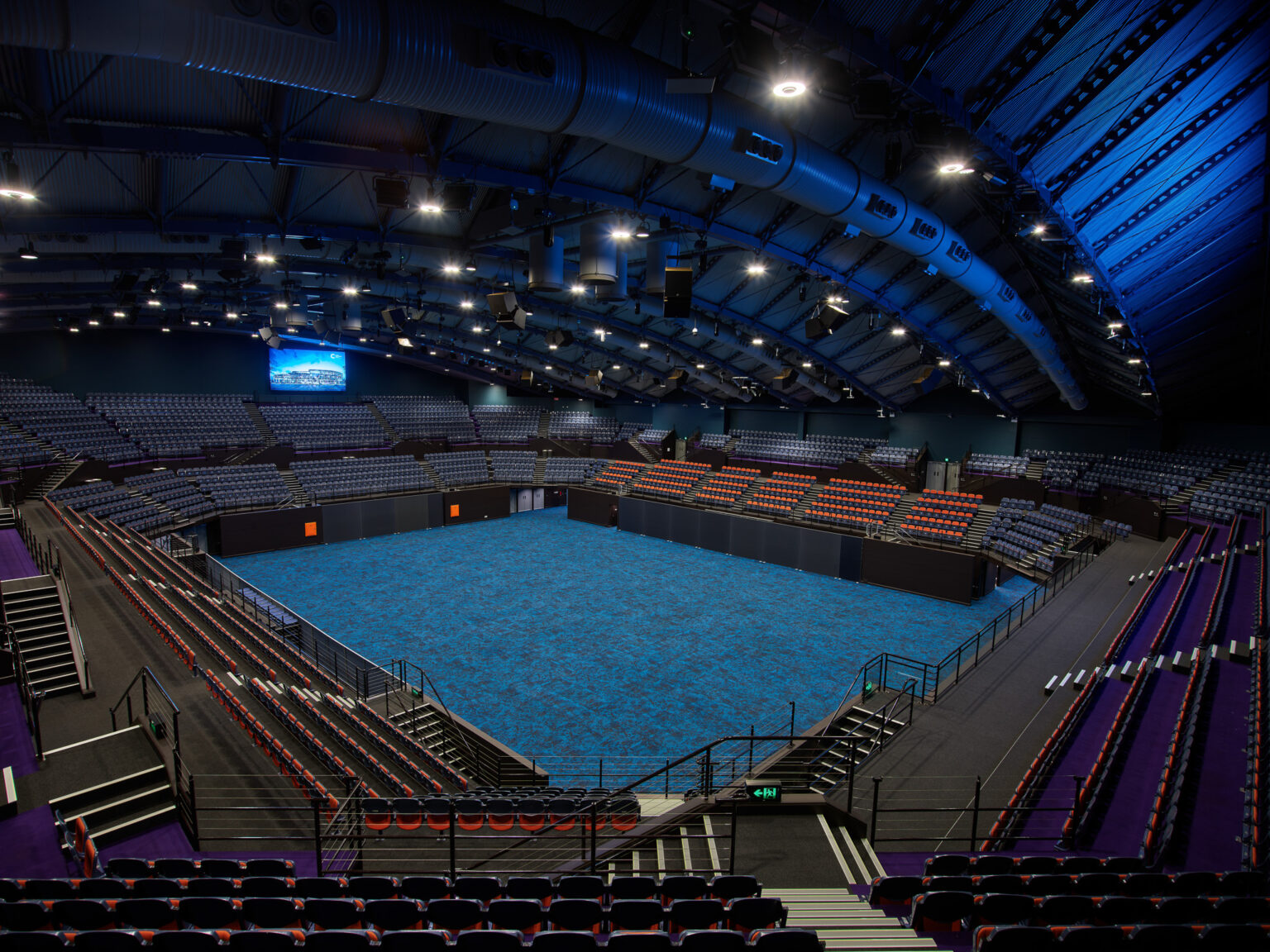 The Cairns Convention Centre Arena is a spacious indoor venue with a large, open floor covered in blue carpet. It is surrounded by multiple tiers of red and black seating. The ceiling has a curved, industrial design with exposed ducts and numerous lights providing bright illumination. A large screen is mounted on one side of the arena, displaying an image or video. The arena is empty, highlighting its modern architecture and substantial seating capacity.