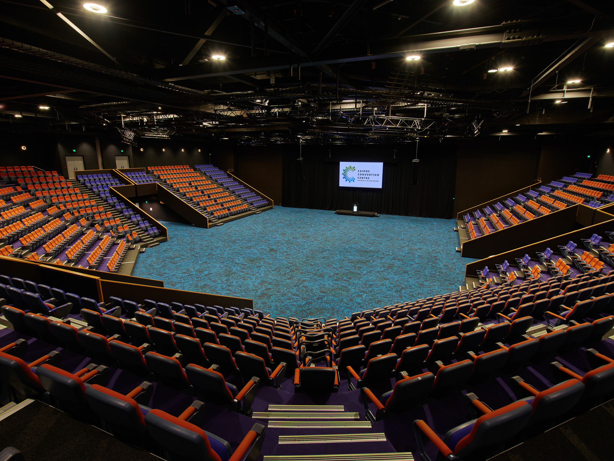 A spacious auditorium with tiered seating arranged in a semi-circular layout around a central stage. The seats are upholstered in blue and orange fabric, creating a vibrant contrast. The stage floor is covered in blue carpet, and a large screen at the back displays the 'Cairns Convention Centre' logo. The ceiling is equipped with lighting fixtures and rigging equipment, enhancing the venue's professional presentation setup