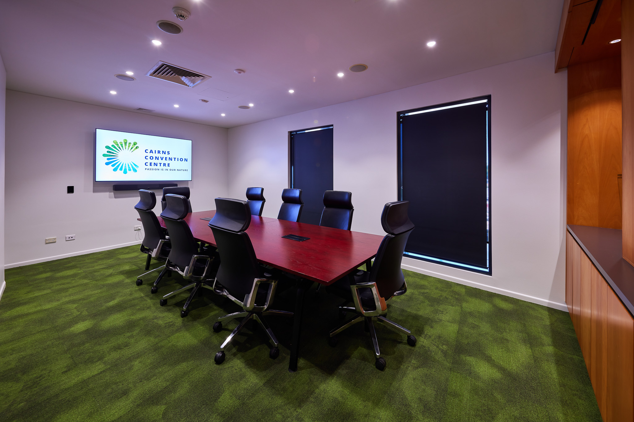 A modern meeting room at the Cairns Convention Centre, labeled as Business Centre Meeting Room 1. The room features a large rectangular table with a glossy red surface surrounded by eight black office chairs on wheels. The floor is covered with green carpet, and the walls are white. On one wall, there is a large flat-screen television displaying an image of a cityscape. Two windows with closed blinds are visible on another wall, and there is recessed lighting in the ceiling providing ample illumination. A small section of cabinetry with a countertop is present in one corner of the room