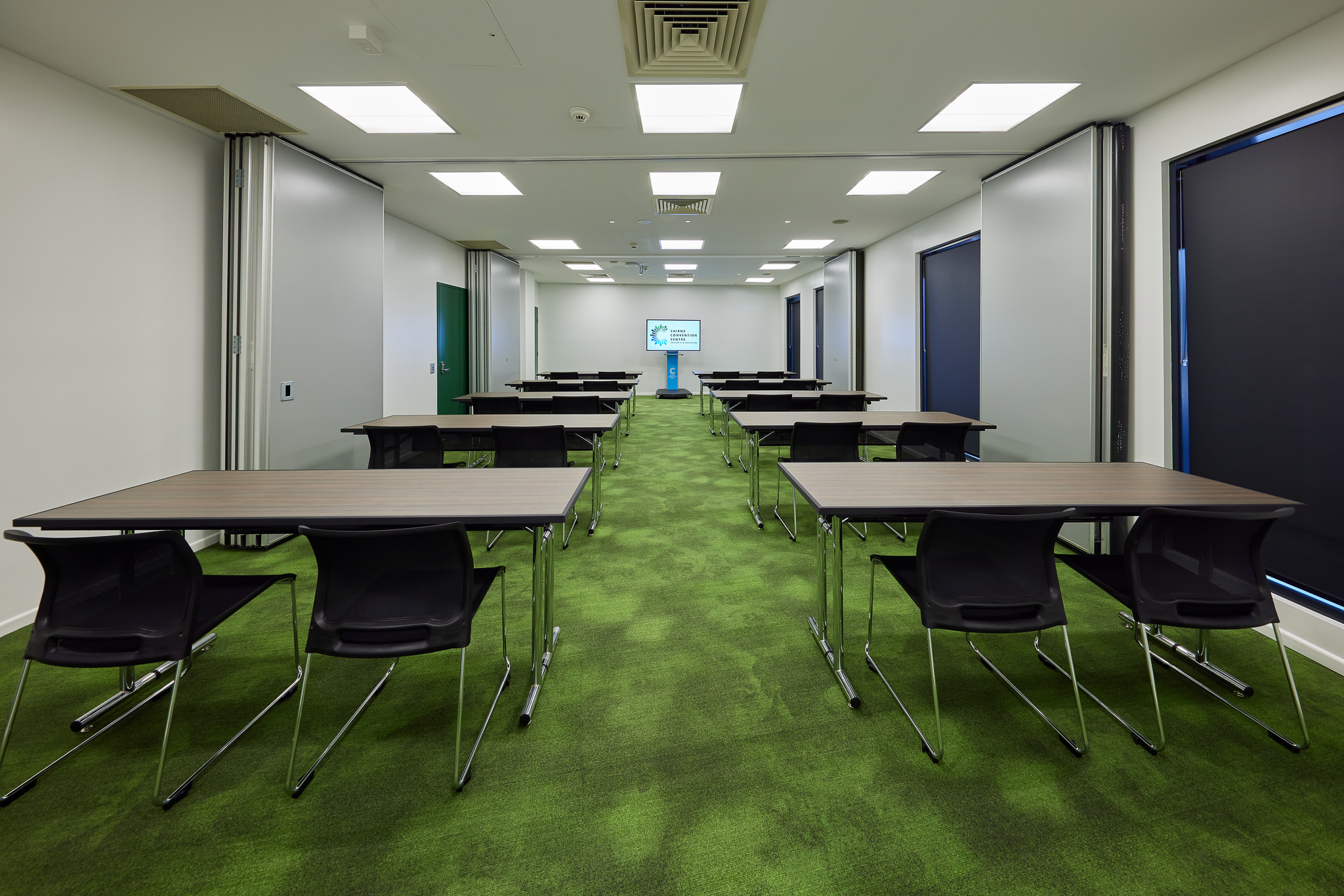 A business centre room at the Cairns Convention Centre, configured for presentations or training sessions. The space features multiple rectangular tables arranged in rows, each paired with black chairs facing a large screen at the front. The floor is covered in green carpet, and the room is well-lit with ceiling lights. Windows along one side have dark blinds partially drawn, creating a professional and focused atmosphere.