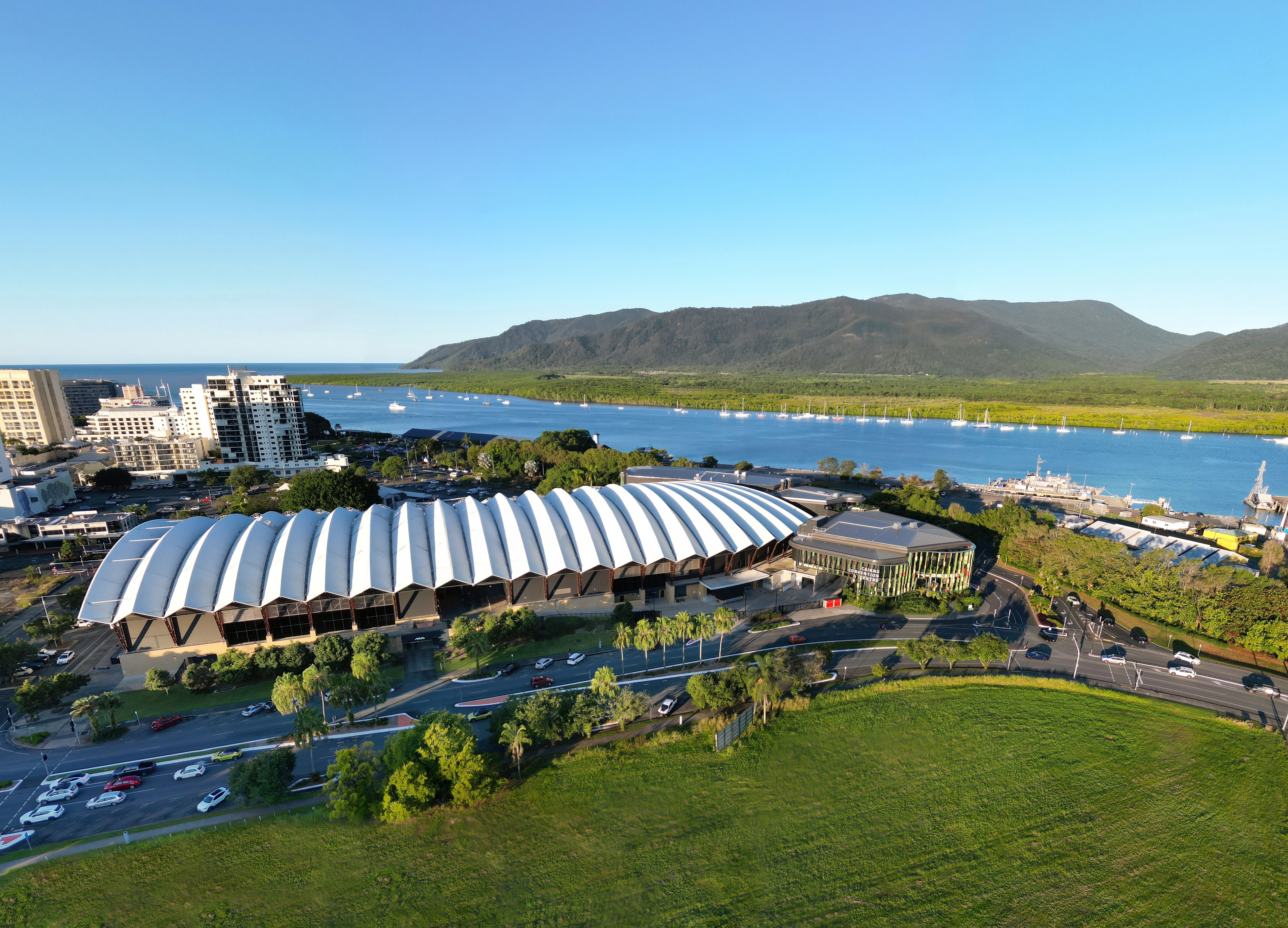 Aerial view of the Cairns Convention Centre with its distinctive white, wavy roof, surrounded by urban buildings and green spaces. In the background, the Trinity Inlet is visible with boats docked along the shore, and beyond it, the lush Wet Tropics Rainforest and distant mountains under a clear blue sky