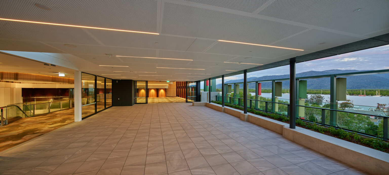 Image of the modern Trinity Terrace with tiled flooring, recessed ceiling lights, glass walls on one side, and open views of greenery and distant mountains under a cloudy sky.