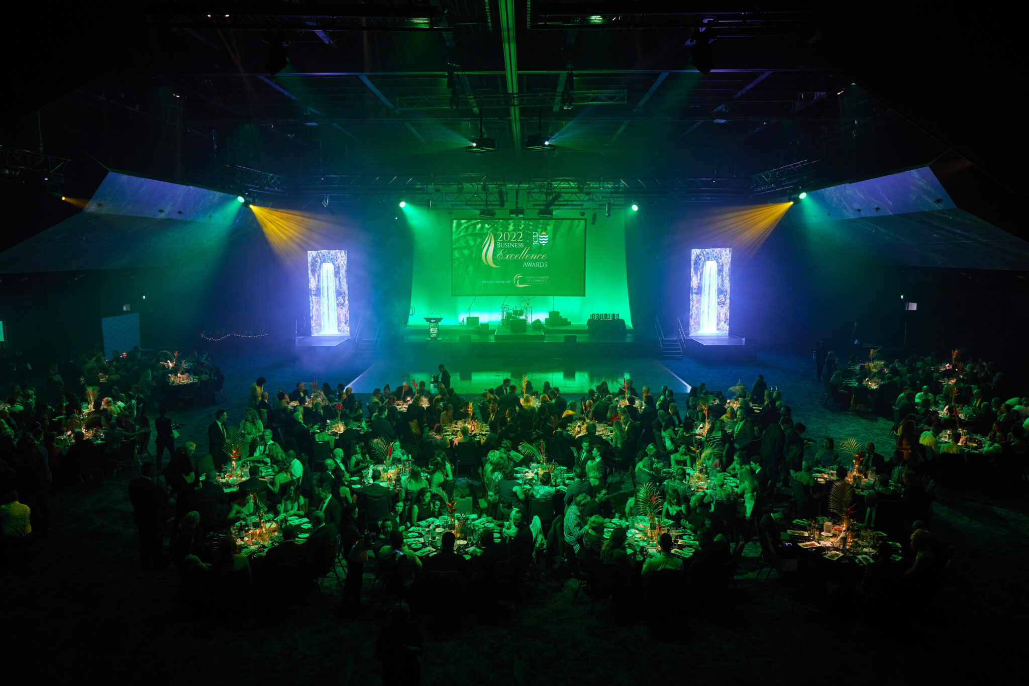 A gala dinner event held in the auditorium of the Cairns Convention Centre. The space is dimly lit with green and yellow lighting, creating an elegant ambiance. Round tables with floral centerpieces fill the room, surrounded by guests in formal attire. A stage at the front features a large screen displaying '2022 Gala Dinner,' flanked by two tall vertical screens with decorative visuals. The setting conveys a sophisticated and celebratory atmosphere