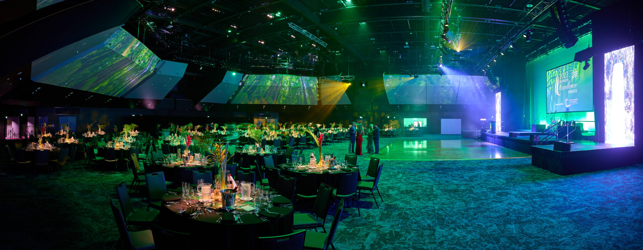 The interior of an auditorium at the Cairns Convention Centre set up for an event. Round tables with black tablecloths are decorated with floral centerpieces and lit candles. Multiple projection screens display green and blue visuals, while stage lighting in green and yellow hues creates a vibrant atmosphere. The stage, visible on the right, is equipped with instruments and microphones, indicating preparations for a performance or presentation. Several people are standing near the center of the room, contributing to the lively ambiance