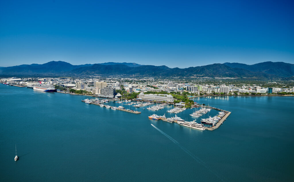 Aerial view of Cairns, a city nestled between the Wet Tropics Rainforest and the Great Barrier Reef. The image shows a sprawling urban area with numerous buildings, roads, and green spaces. The coastline features a marina with several boats docked and others sailing in the water. In the heart of the city is the Cairns Convention Centre, highlighted as a central hub for business events and conferences. The surrounding landscape includes lush greenery from the rainforest and distant mountain ranges under a clear blue sky. This image beautifully captures Cairns' unique position between natural wonders and its role as a significant destination for business events.