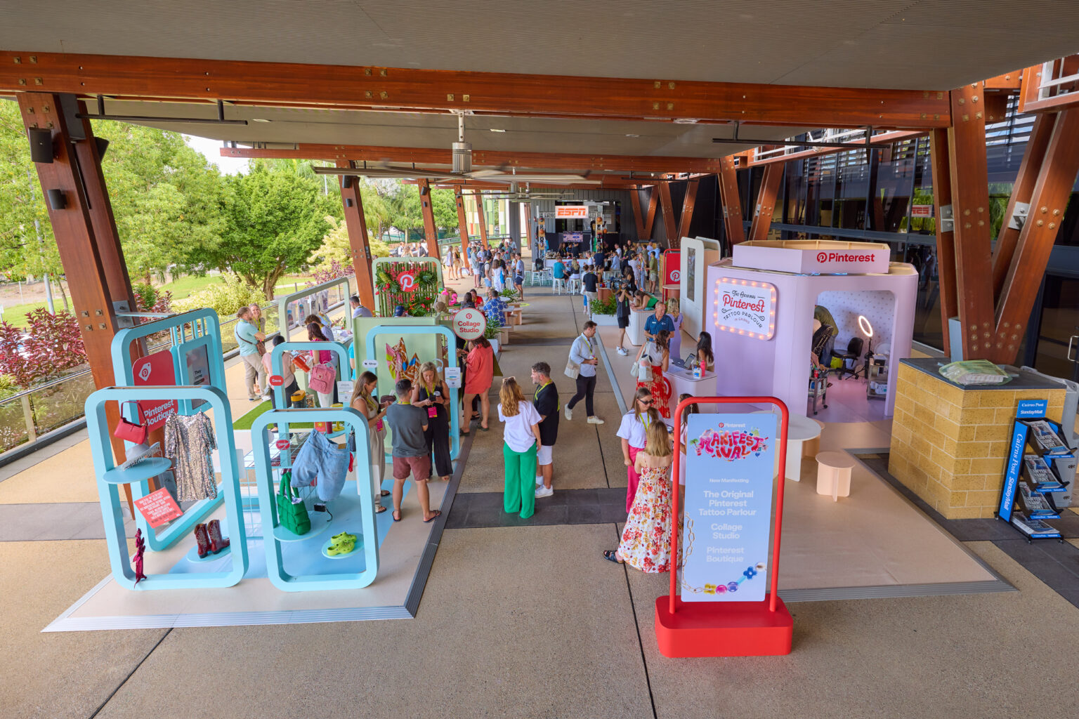 An outdoor exhibition event on the City Terrace at the Cairns Convention Centre. The space features various colorful booths and displays under a large canopy structure supported by wooden beams. Attendees are seen walking around and engaging with the exhibits. One booth prominently displays a sign reading 'Pinterest Presents' with a list of activities. Lush greenery is visible in the background, enhancing the open and vibrant atmosphere