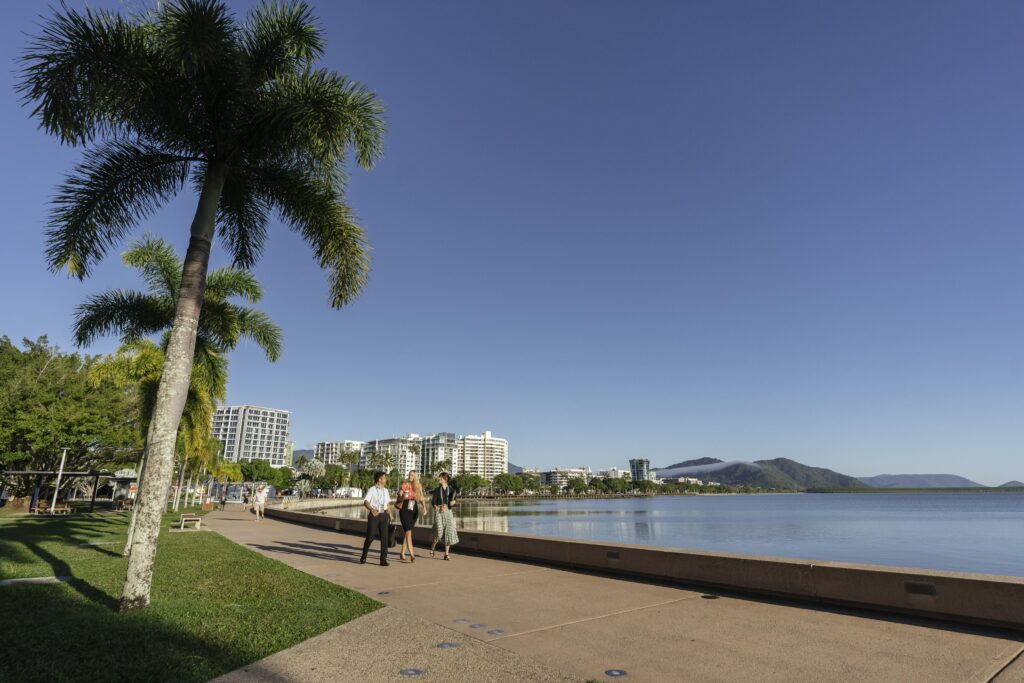 Scenic view along the Cairns Esplanade featuring palm trees lining a paved walkway with people strolling. To the right, a calm body of water reflects the clear blue sky. In the background, modern buildings and distant mountains complete the tropical landscape.