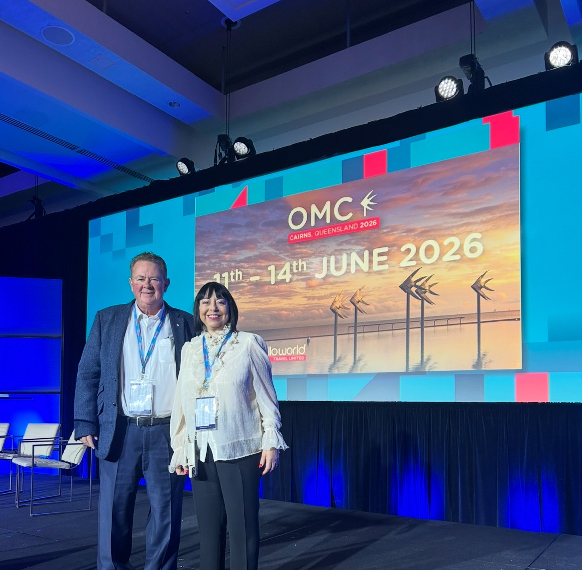 Andrew Burnes, AO and Cinzia Burnes (HLO) announcing the destination at the 2025 Helloworld Travel Owner Manager Conference in Vancouver. The image shows the two individuals standing on a stage with a large screen behind them displaying 'OMC Cairns Queensland 2026' and the dates '11th - 14th June 2026