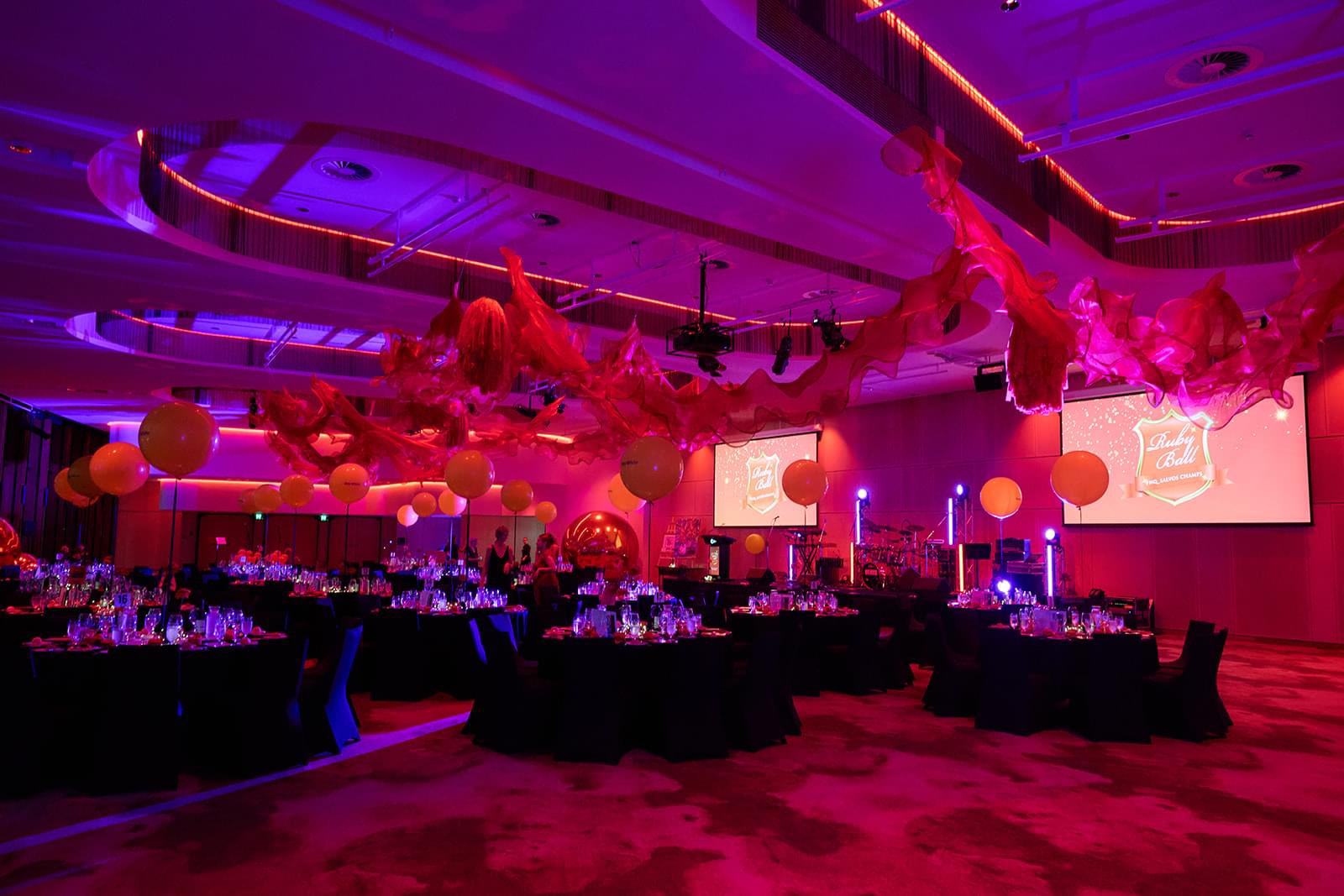 A large, elegantly decorated banquet hall with purple and pink lighting in the Trinity Room at Cairns Convention Centre. The ceiling is adorned with flowing red fabric and large yellow balloons. Round tables covered in black tablecloths are set up throughout the room, each adorned with glassware and centerpieces. Two large projection screens display a logo that reads 'Ruby Ball' in white text on a red background. The atmosphere is festive and vibrant, suggesting an event or celebration is taking place