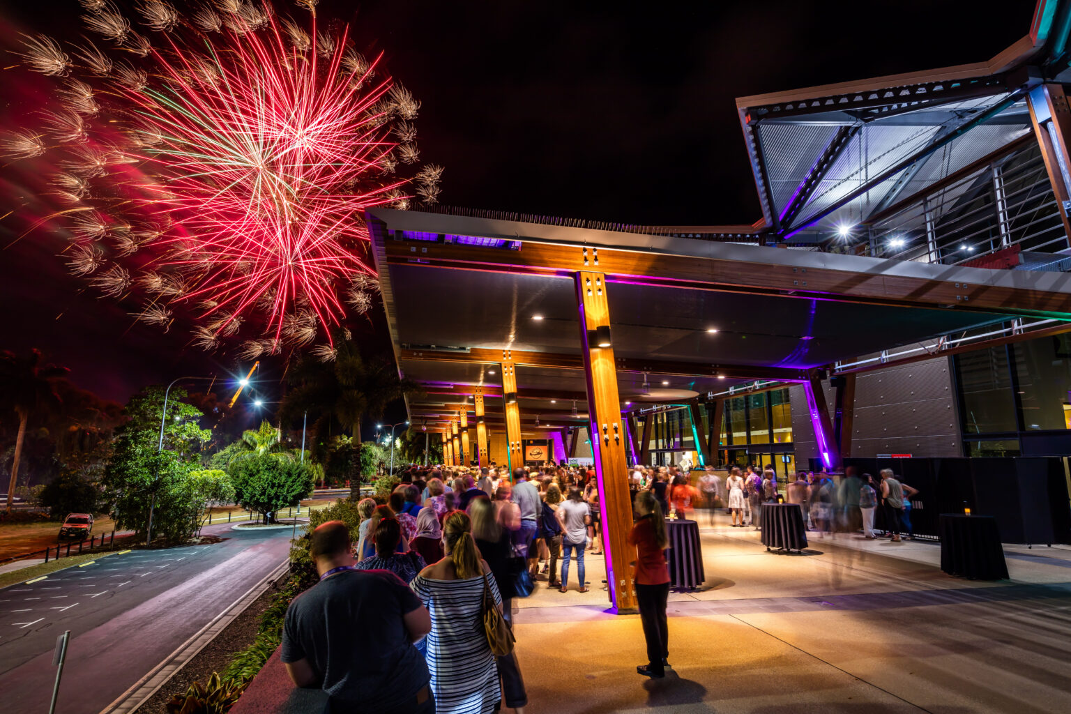 A festive night scene at the City Terrace of the Cairns Convention Centre, with a crowd of people gathered outside a modern building featuring large glass windows. The area is illuminated by colorful lights, and vibrant red fireworks light up the sky in the background, creating a celebratory atmosphere