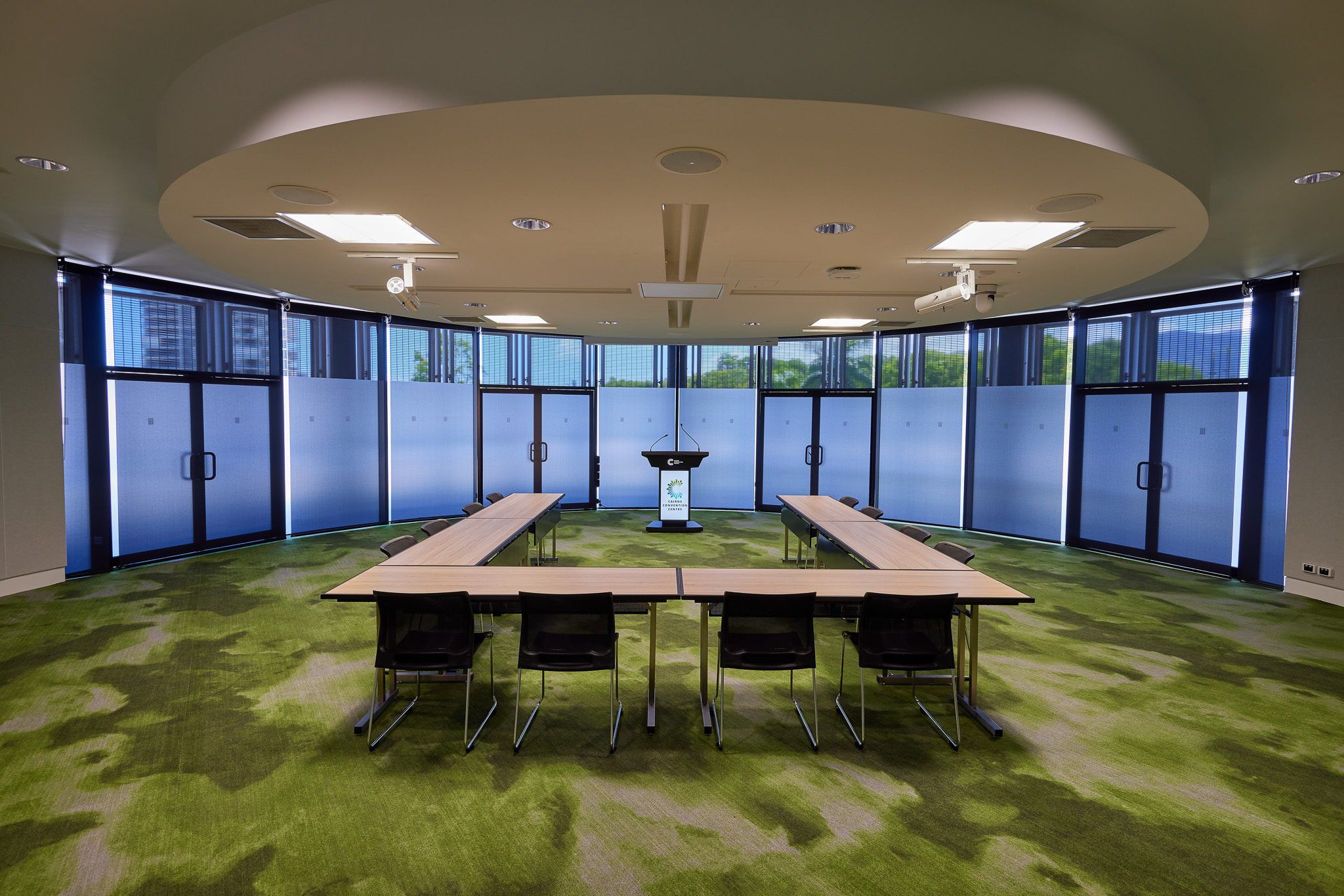 A well-lit meeting room at the Cairns Convention Centre featuring a U-shaped arrangement of tables and chairs. A podium is positioned at the center of the U-shape, suitable for presentations or speeches. The room has large windows with frosted glass on the lower half, allowing natural light while maintaining privacy. The ceiling includes recessed lighting and two ceiling-mounted projectors. The floor is covered in green carpet