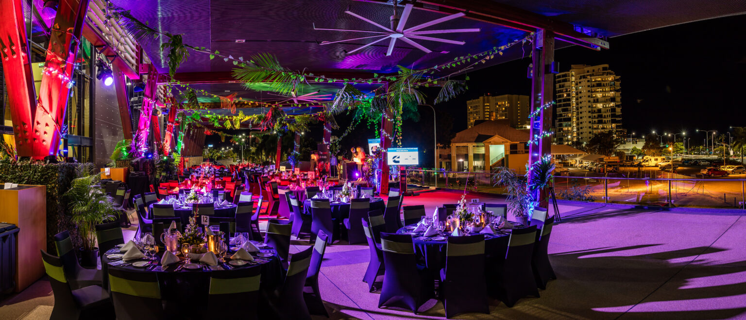 An outdoor evening event setup on the City Terrace at the Cairns Convention Centre. Round tables with black tablecloths are arranged throughout the space, each decorated with candles, glasses, and napkins. The area is lit with colorful lighting in shades of purple, red, and green. Palm trees and plants enhance the tropical ambiance, while buildings and a parking lot are visible in the background under the night sky