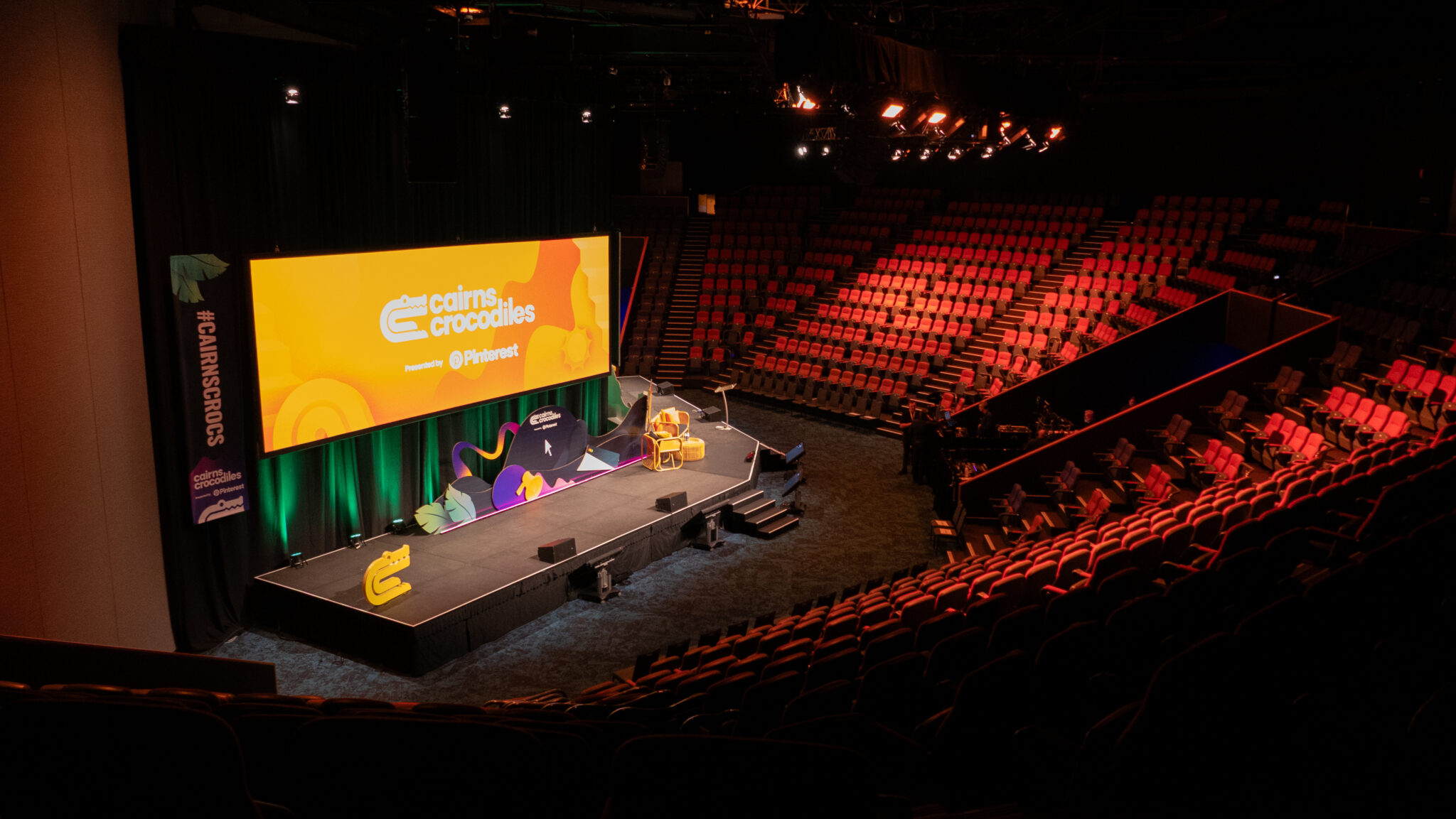 An auditorium with a large stage at the front, featuring a bright yellow screen displaying the text 'Cairns Crocodiles' and '@Pinterest.' Two chairs, including one orange, are placed on the stage. The seating area consists of red and black seats arranged in a semi-circular pattern. The auditorium is dimly lit, with spotlights focused on the stage.