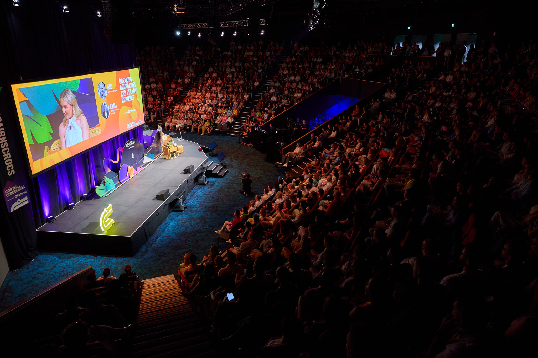 A large auditorium filled with an audience seated in rows, facing a stage at the front. The stage features a large screen displaying colorful graphics and text. The room is dimly lit, with the stage area brightly illuminated, creating a focal point for the presentation or event taking place