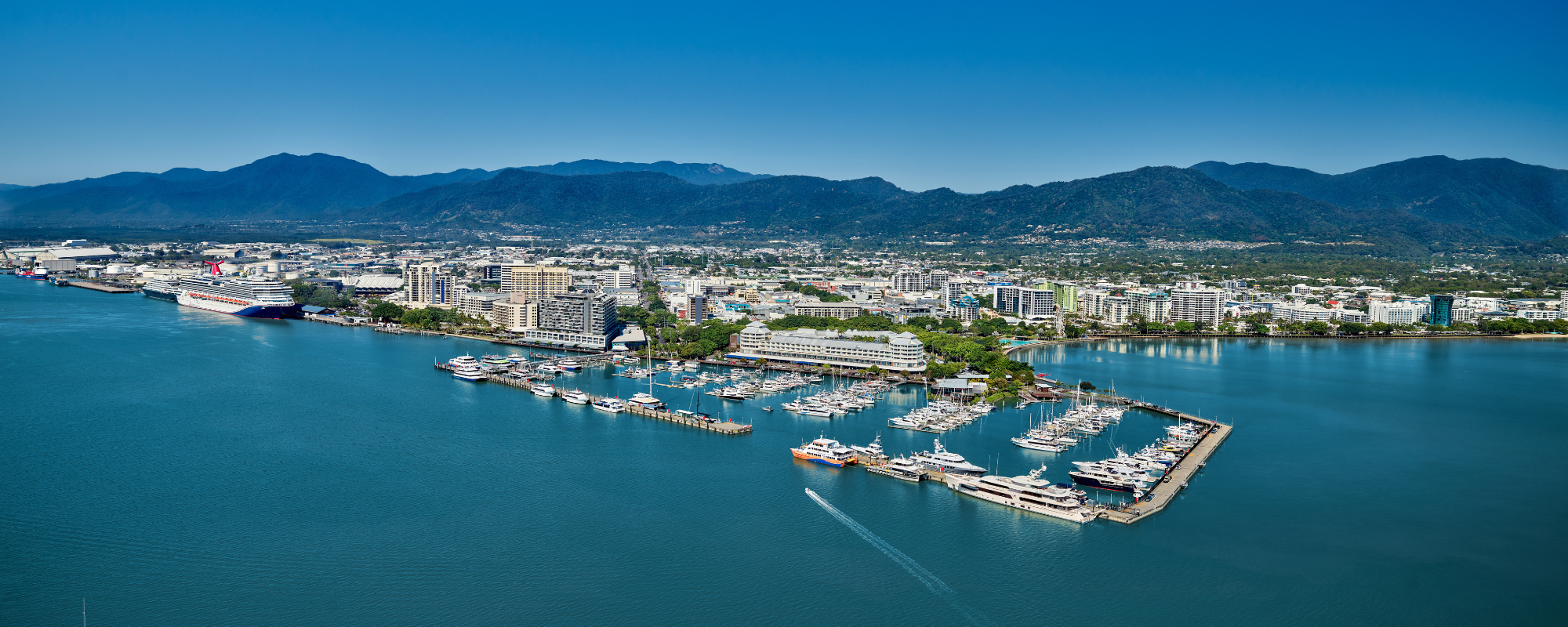 Aerial view of Cairns, a city nestled between the Wet Tropics Rainforest and the Great Barrier Reef. The image shows a sprawling urban area with numerous buildings, roads, and green spaces. The coastline features a marina with several boats docked and others sailing in the water. In the heart of the city is the Cairns Convention Centre, highlighted as a central hub for business events and conferences. The surrounding landscape includes lush greenery from the rainforest and distant mountain ranges under a clear blue sky. This image beautifully captures Cairns' unique position between natural wonders and its role as a significant destination for business events.