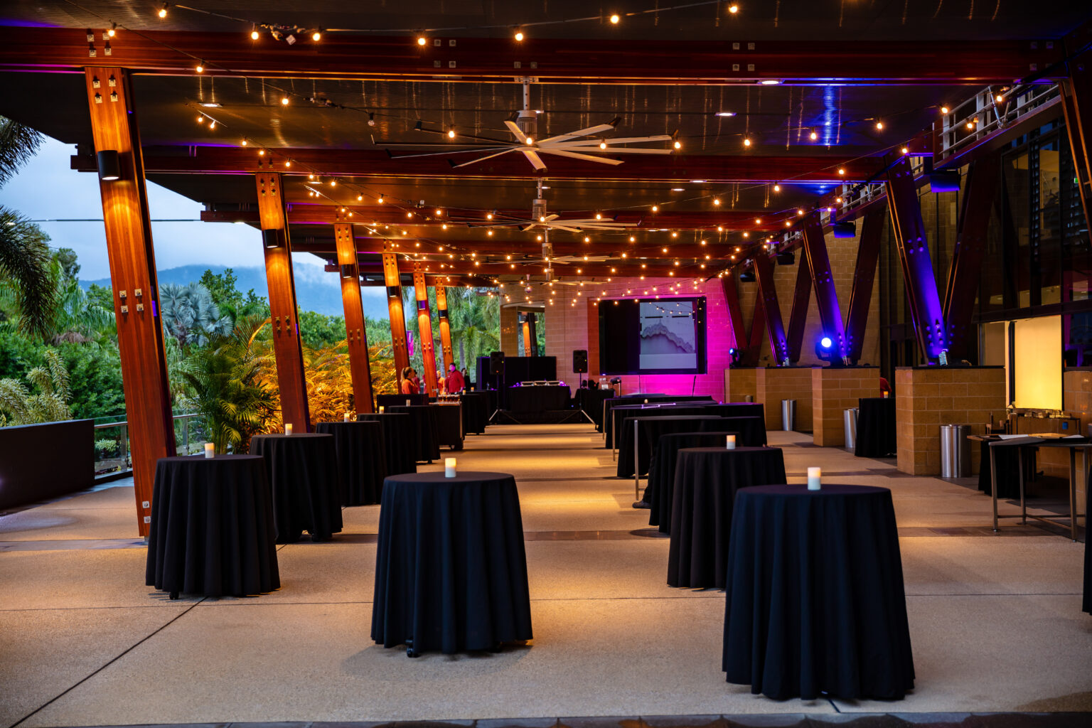 The City Terrace at the Cairns Convention Centre set up for an exhibition and cocktail function. The covered outdoor space features string lights hanging from the ceiling, creating a warm and inviting ambiance. Round cocktail tables with black tablecloths and candle centerpieces are arranged throughout the area. Large wooden beams support the structure, and the terrace overlooks lush greenery. A stage with lighting and audio equipment is visible in the background, prepared for presentations or performances