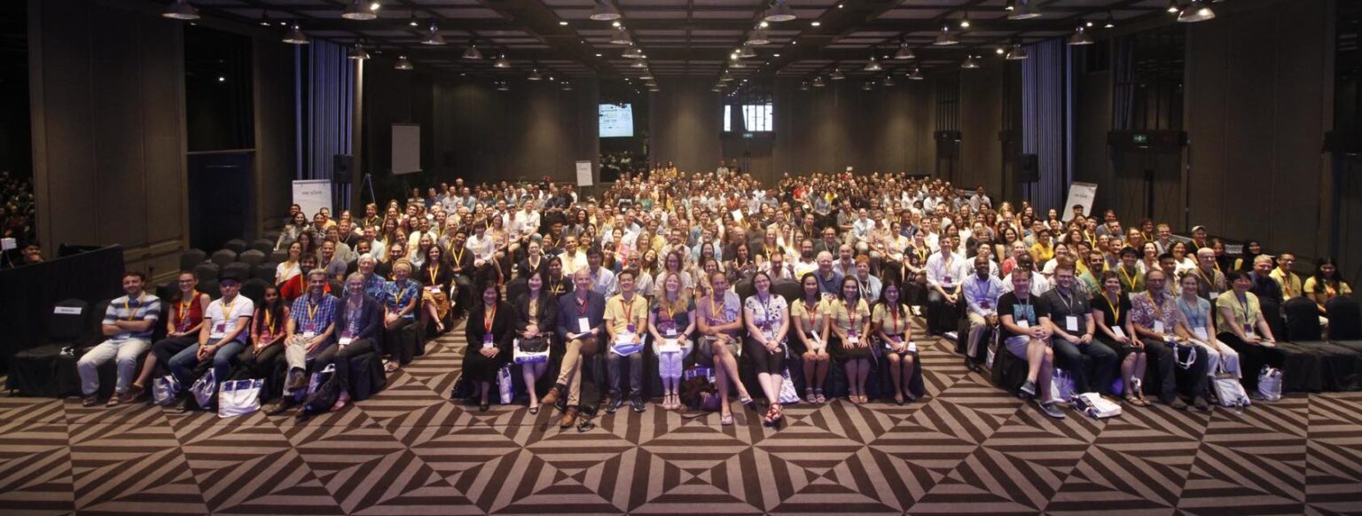 A large group of people seated in rows from the International Bat Research Conference inside a spacious conference hall with geometric patterned carpet. The attendees are facing the camera, and the hall is well-lit with ceiling lights. There are projection screens visible in the background