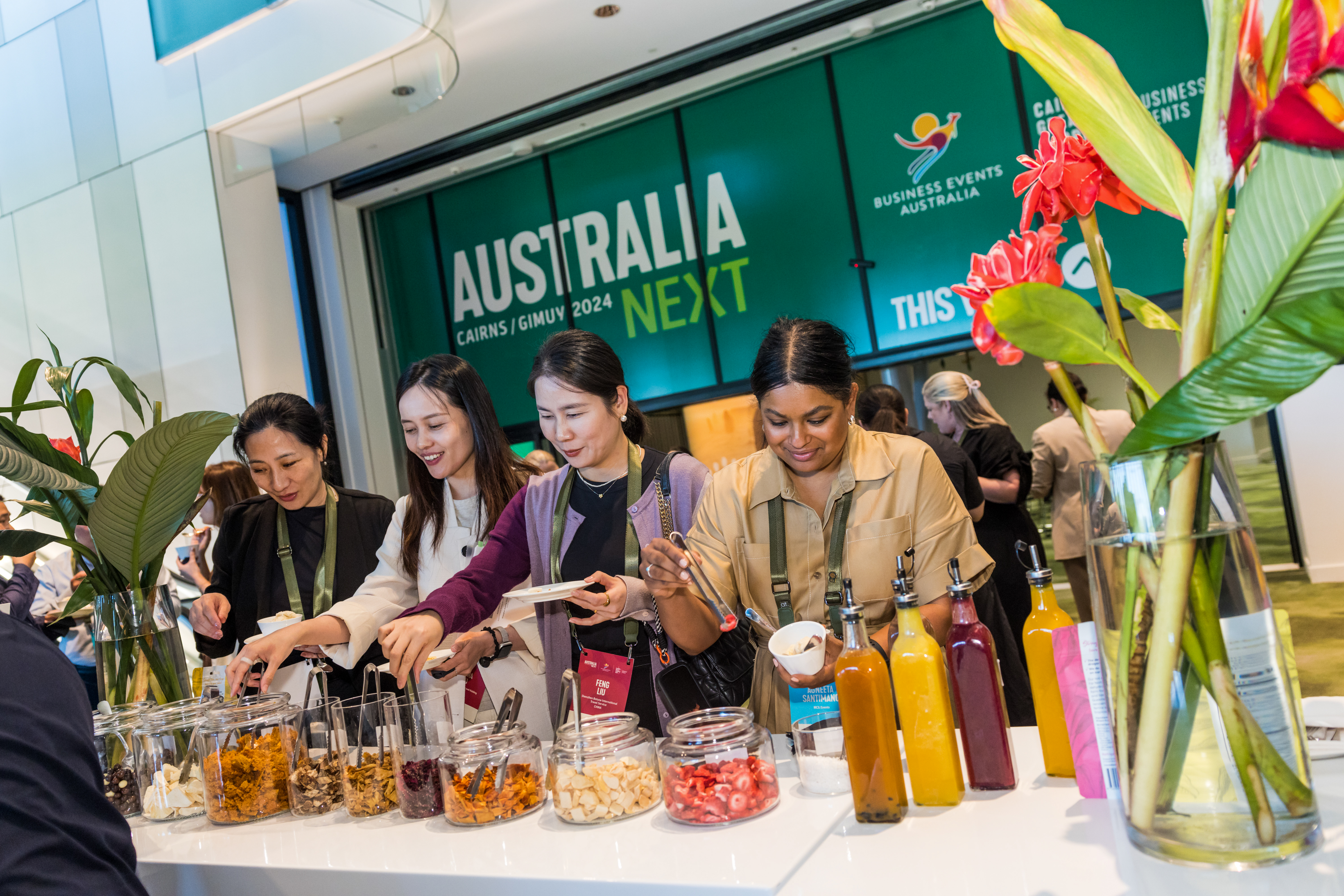 group of people standing at a buffet table displaying various jars and bottles of food items, with decorative plants and flowers. In the background, a large sign reads 'AUSTRALIA NEXT' with additional text 'CAIRNS/GIMUY 2024' and 'BUSINESS EVENTS AUSTRALIA.