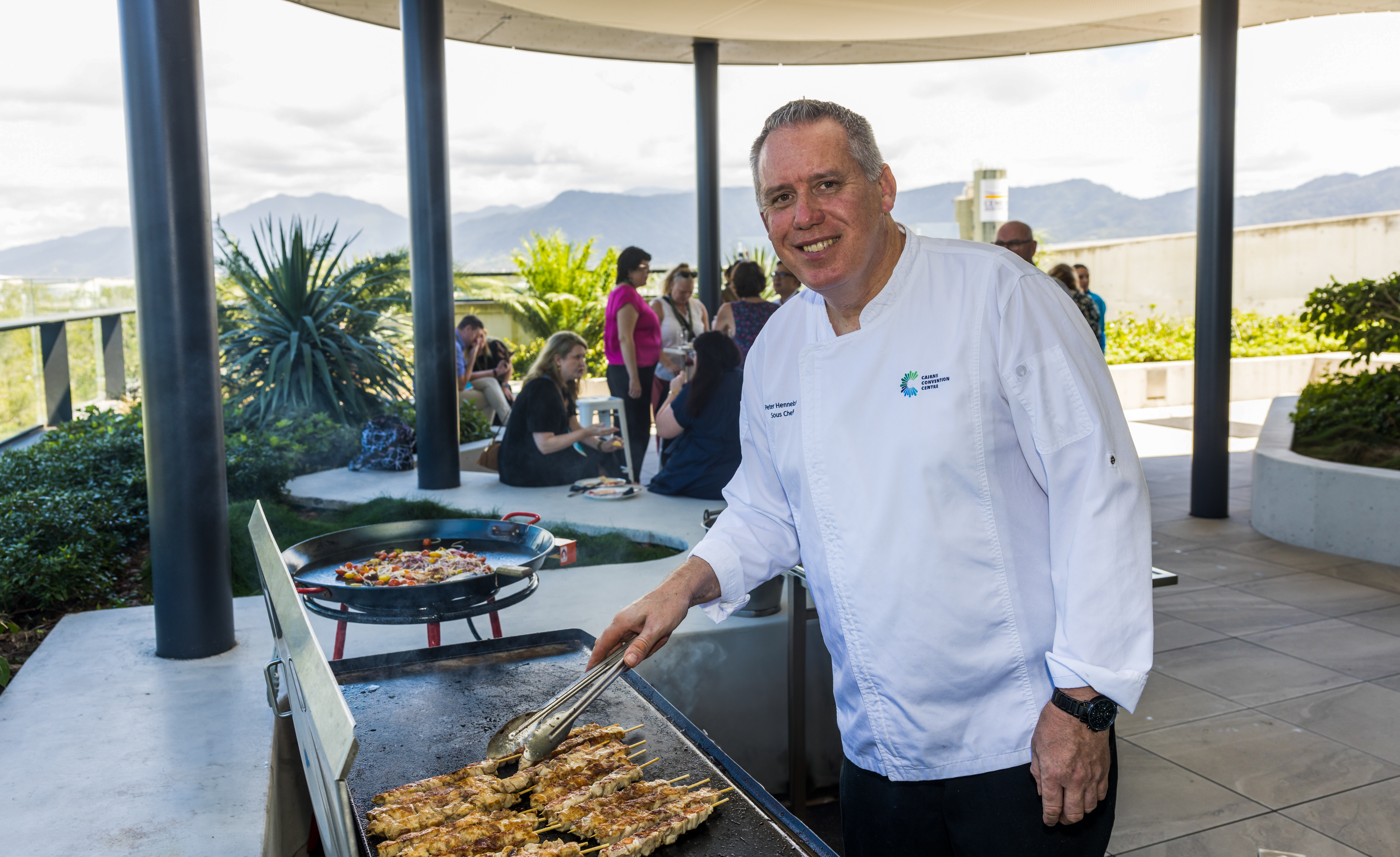 A chef in a white uniform grilling food on an outdoor flat-top grill, with their face obscured. In the background, people are gathered around a circular seating area, surrounded by greenery and distant mountains. A second cooking station with a large pan of food and a can of cooking spray is visible nearby.