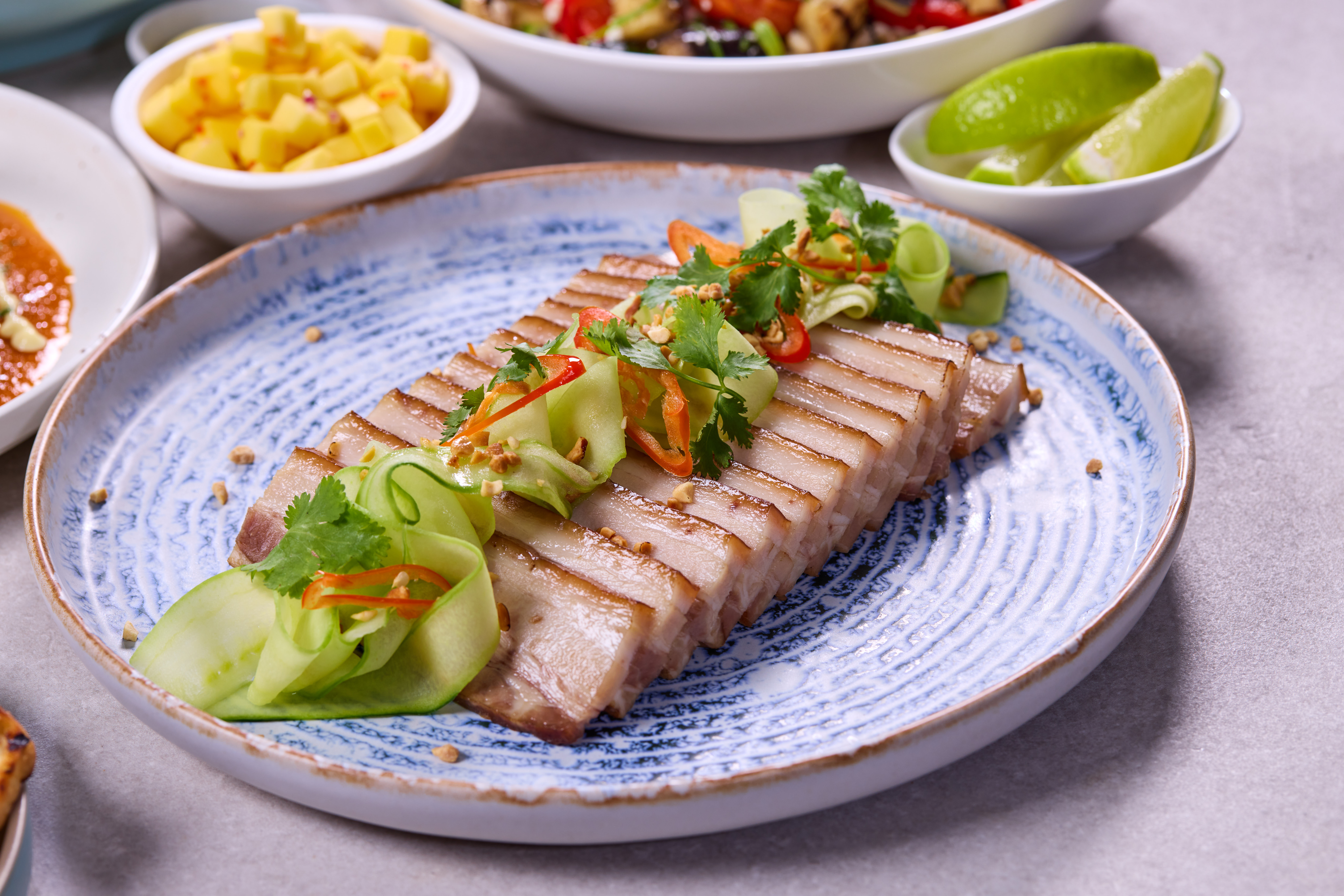 A plate of pork belly slices garnished with cucumber ribbons, red chili slices, and cilantro, served on a blue and white ceramic plate. In the background, small bowls hold lime wedges, diced mango, and a colorful rice noodle salad