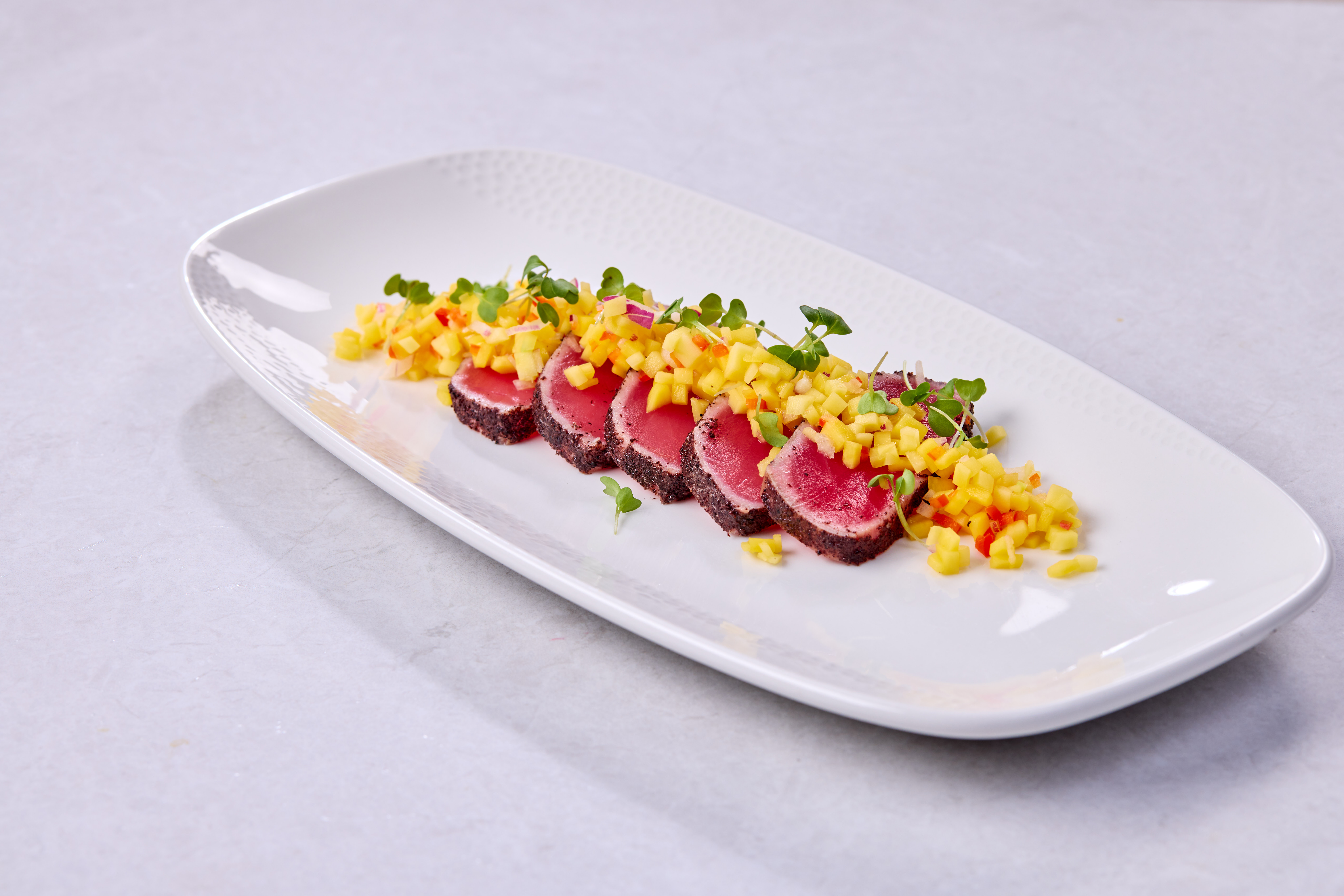 Slices of seared tuna with pink centers and dark seared edges, topped with mang and microgreens, served on a white plate