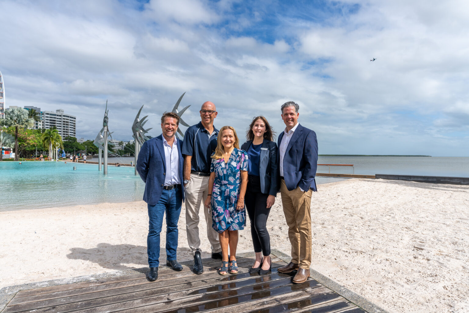 Five professionally dressed individuals stand on a wooden platform by a tropical beach, with clear blue water, sculptures, and palm trees in the background. The group appears to be promoting the CAPA Conference, set against a scenic coastal backdrop with partly cloudy skies and distant buildings.