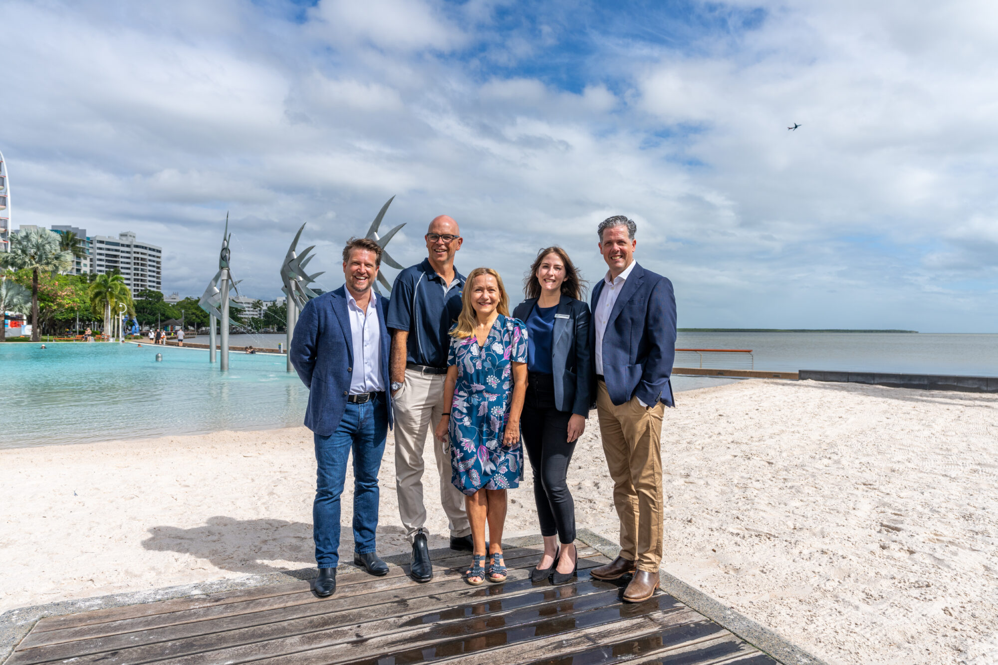 Five professionally dressed individuals stand on a wooden platform by a tropical beach, with clear blue water, sculptures, and palm trees in the background. The group appears to be promoting the CAPA Conference, set against a scenic coastal backdrop with partly cloudy skies and distant buildings.
