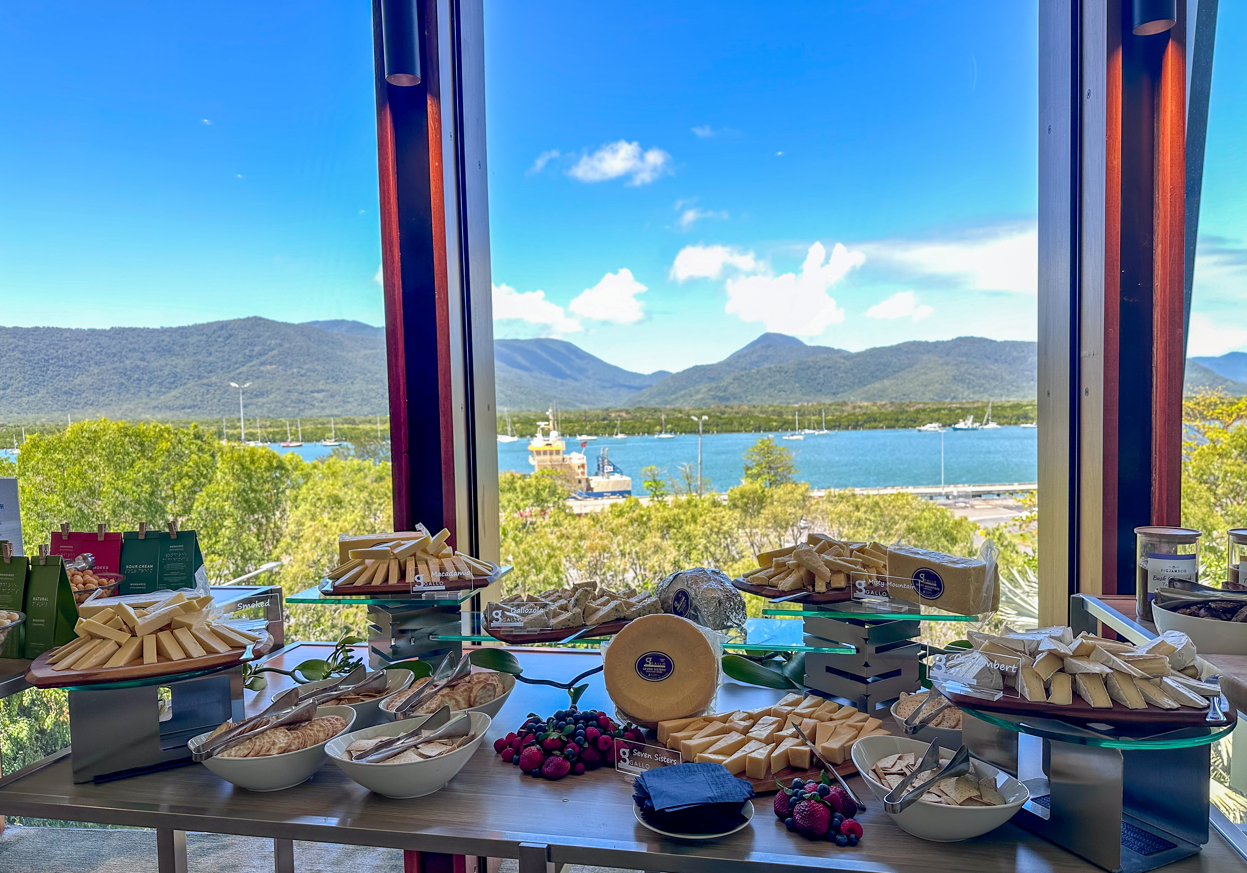 A buffet table arranged with assorted cheeses, crackers, and fruits, set in front of large windows overlooking a scenic body of water with boats and a mountainous landscape. The sky is partly cloudy, creating a bright and inviting atmosphere