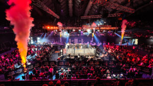 The image shows a large indoor boxing arena filled with spectators. In the centre, there is a boxing ring with several people inside and around it, including fighters and officials. Bright lights illuminate the ring, and there are smoke effects on both sides of the arena. The audience is seated in rows surrounding the ring, and there are banners with "NO LIMIT BOXING" logos around the venue.