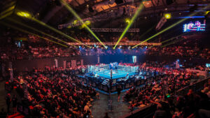 A large indoor arena filled with spectators watching a boxing match. The ring is illuminated by bright yellow spotlights, and there are multiple rows of seating surrounding the ring. The ceiling features industrial lighting and rigging equipment. The audience is seated in tiered sections, with many people standing around the perimeter of the ring area. There are "No Limit" banners displayed around the ring and on the walls of the arena.