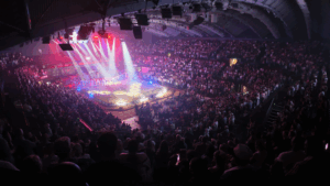 A large indoor arena filled with a crowd of spectators. The central area is illuminated by bright spotlights, revealing a circular stage or ring surrounded by barriers. A group of performers stands in a line within the ring. The ceiling features structural beams and lighting equipment, contributing to the overall atmosphere of the event.