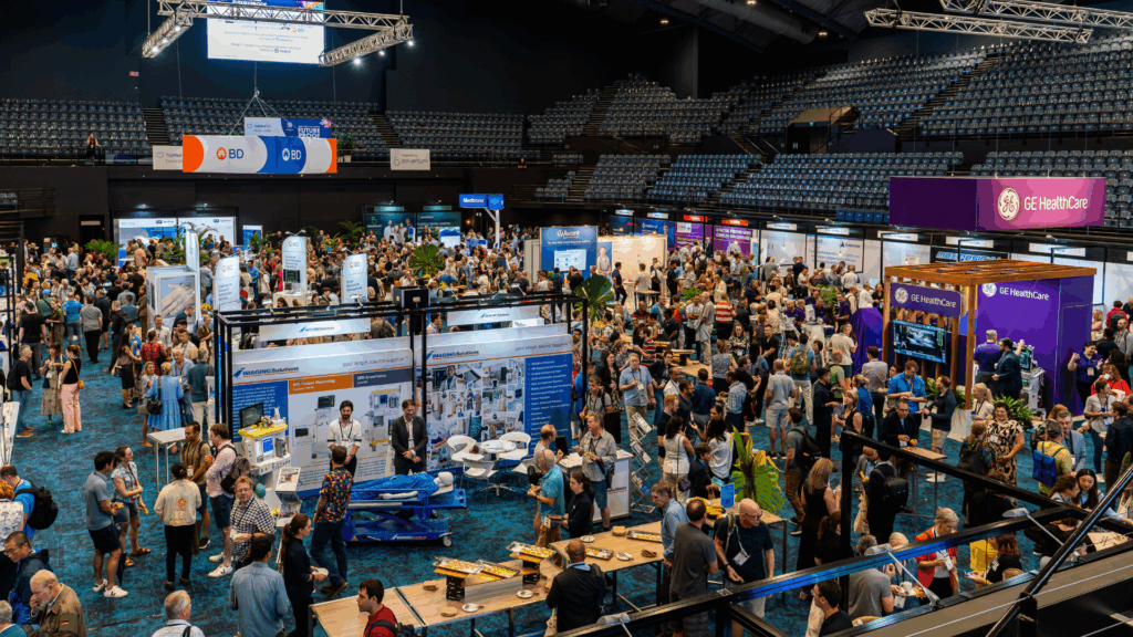 A bustling exhibition taking place in the arena at the Cairns Convention Centre. The space is filled with numerous booths and display panels, with attendees walking around, engaging with exhibitors, and viewing the exhibits. The arena features a high ceiling with lighting rigs and banners from various organizations, including GE Healthcare, contributing to a dynamic and professional exhibition environment