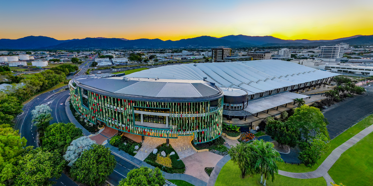 An aerial view of the Cairns Convention Centre at sunset, featuring a curved facade with vertical green, red, and white panels. The Centre is surrounded by landscaped greenery, well-maintained roads, and nearby buildings. In the distance, industrial structures and mountains are visible under a vibrant sky transitioning from yellow to blue