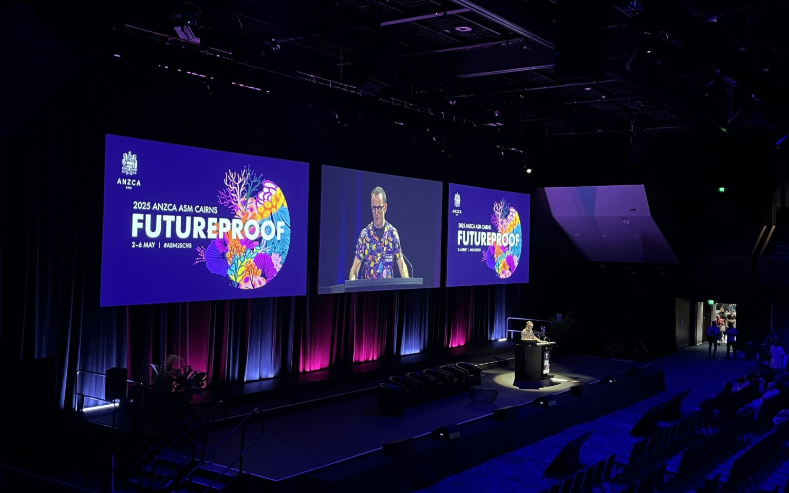Dr Andrew Potter stands at a podium on a stage, delivering a presentation at the 2025 ANZCA ASM Cairns conference. The stage features three large screens displaying the conference's theme "FUTURE PROOF" with colorful coral reef graphics. The event is scheduled for 2–6 May, and the hashtag #ASM25CNS is visible on the screen.