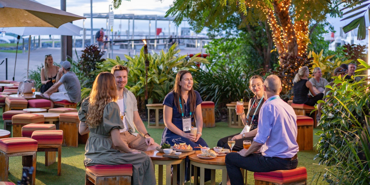 A group of five people are sitting around a circular wooden table in an outdoor setting, enjoying drinks and snacks. The area is decorated with lush greenery, string lights wrapped around a tree, and colorful cushioned stools. Other groups of people can be seen in the background, also seated and conversing. The atmosphere appears relaxed and social.