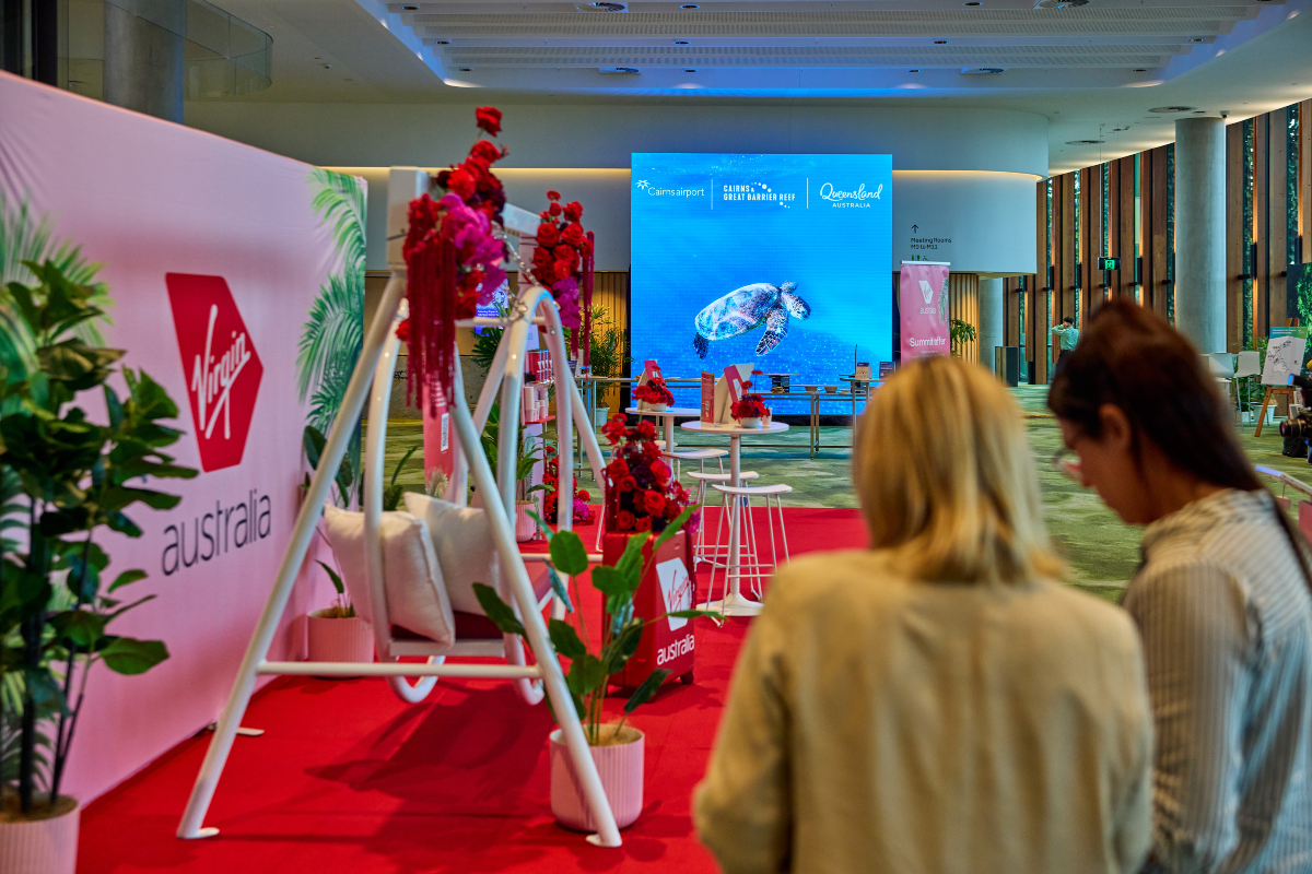 Indoor event setup featuring a Virgin Australia-branded display with a white swing decorated with red flowers and cushions, surrounded by potted plants on a red carpet. In the background, a large digital screen shows an underwater image of a sea turtle with text promoting Queensland. High ceilings, glass walls, and natural light create an open, modern atmosphere.