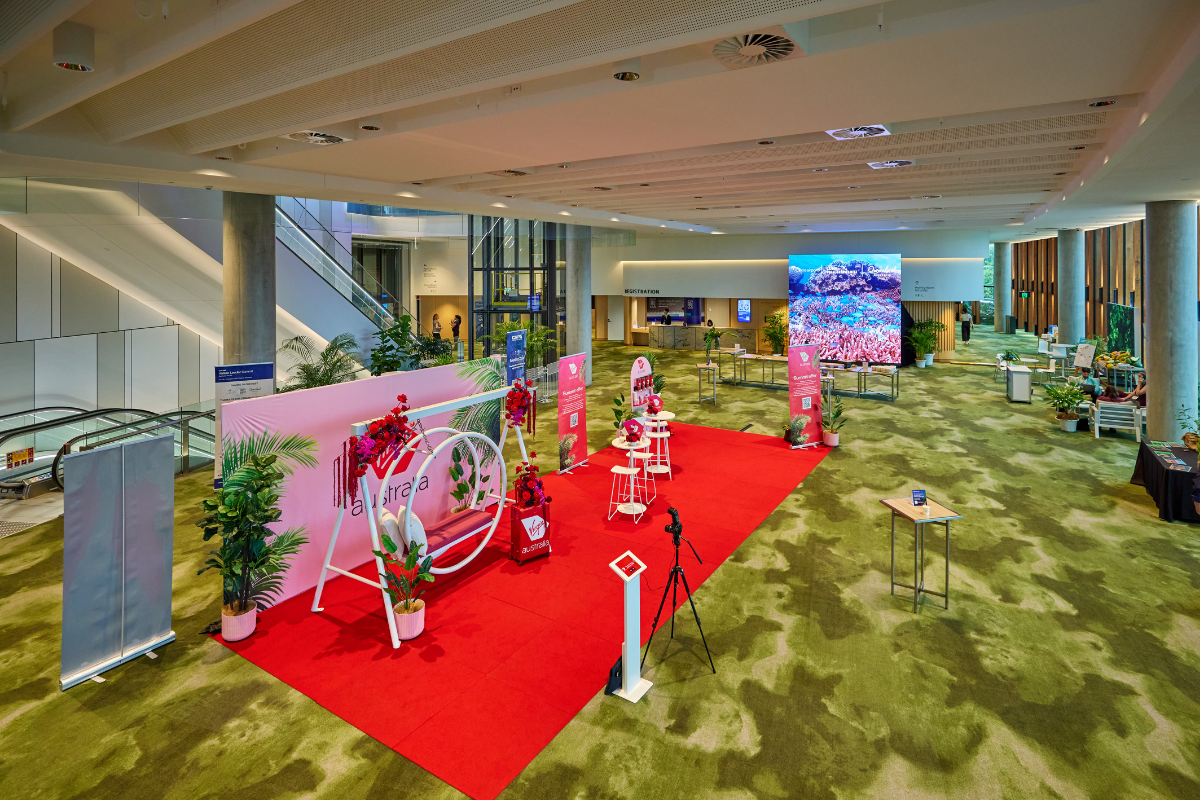 Wide-angle view of the Mezzanine Exhibition event space featuring a Virgin Australia-branded activation area on a bright red carpet. The setup includes a white swing decorated with red flowers, potted green plants, and small tables with stools. A large digital screen in the background displays an image of a vibrant coral reef. The venue has high ceilings, green-patterned carpet, and modern architectural elements such as glass walls, escalators, and concrete columns.